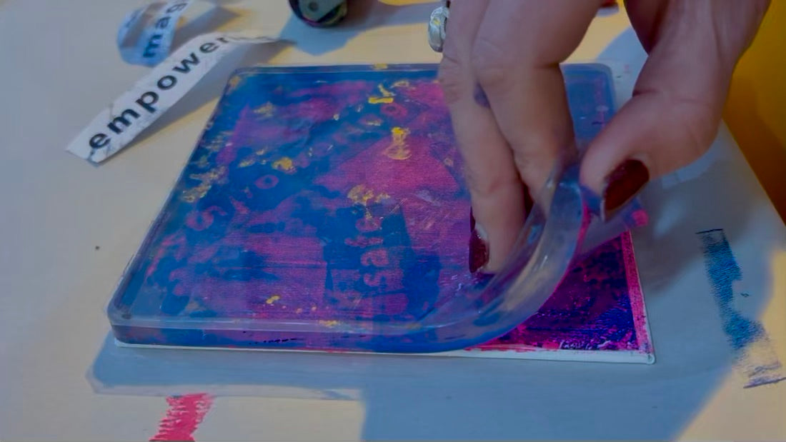A hand peeling back a gel plate to reveal a bright pink and blue print with the words safe and empowering on