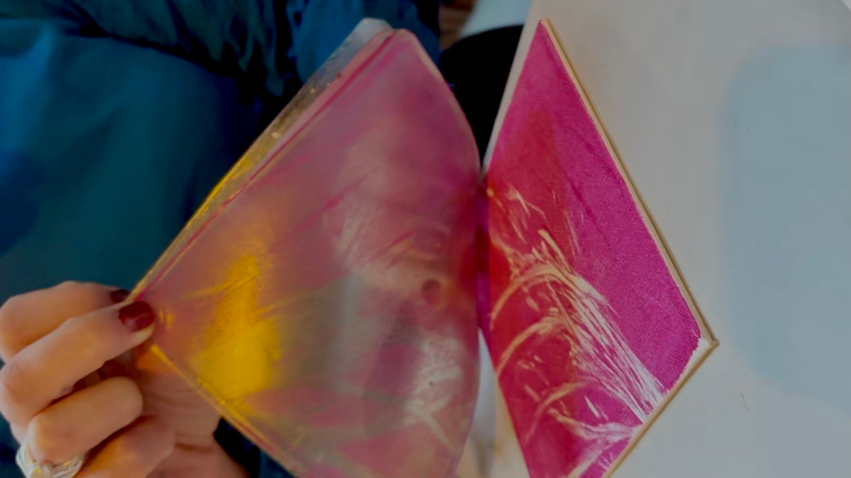 A hand peeling back a gel plate from a square canvas to reveal a bight pink print of some pampas grass