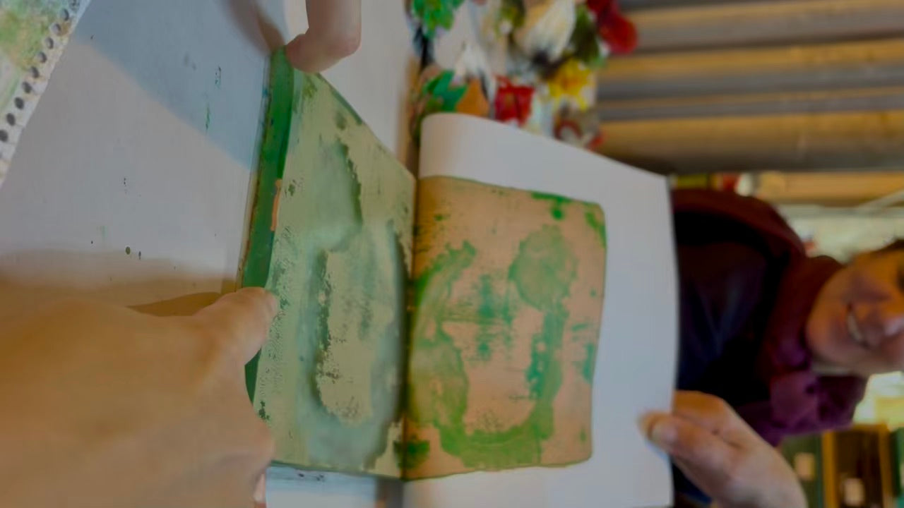 A hand peeling back paper from a gel plate to reveal a green and gold nature inspired 'luck' print in the shape of a horseshoe. In the background are artificial flowers used in the process and a smiling woman.