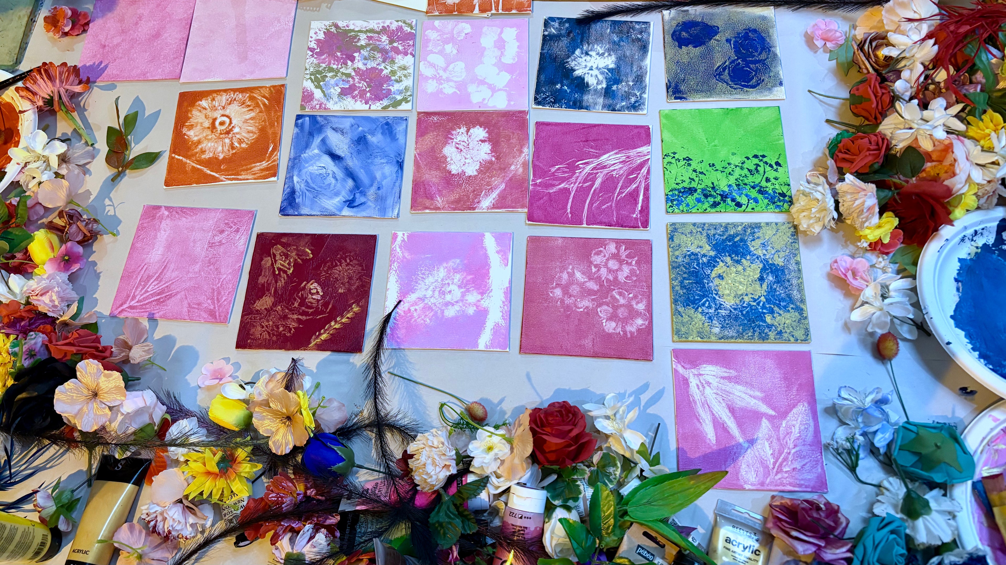 Many brightly painted nature inspired canvases with prints of botanicals on a table surrounded by lots of tubes of paint and artificial flowers