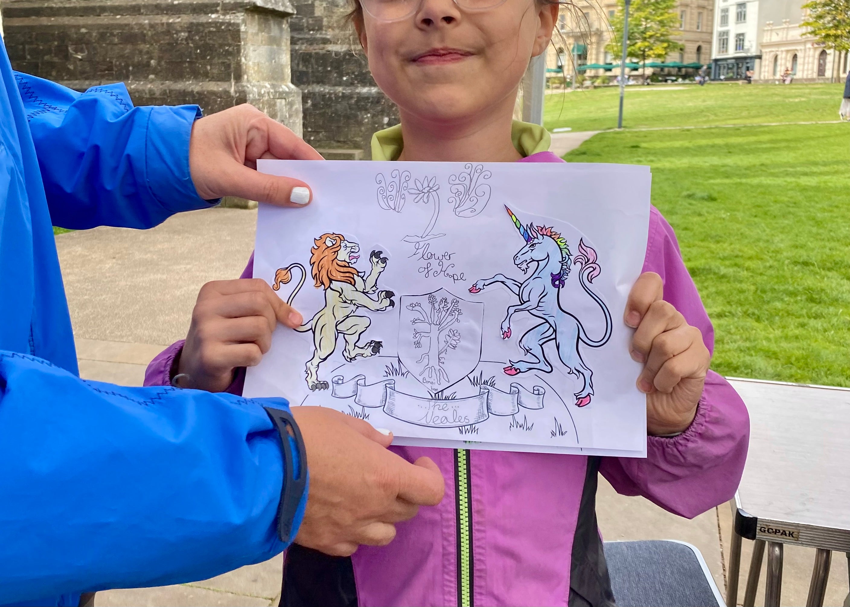 A girl holding up a sheet of paper with a custom crest on. The crest has a unicorn, a lion and other motifs and has been coloured in using colouring pencils. The cathedral wall and green are in the background