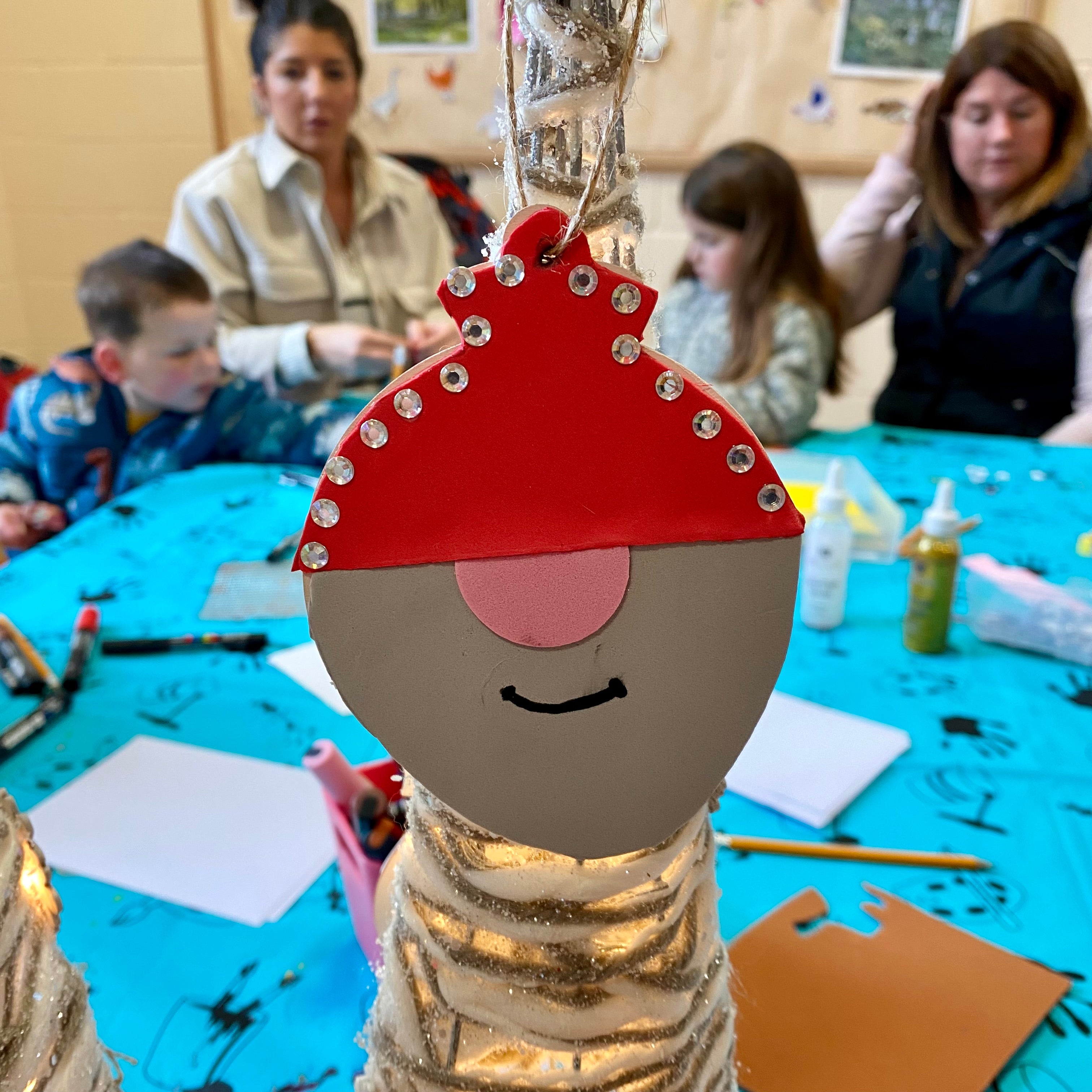 A gnome shaped crafted bauble with sparkly gems on a Christmas tree. In the background is a workshop in full swing with other people sat around a table creating their own custom baubles.