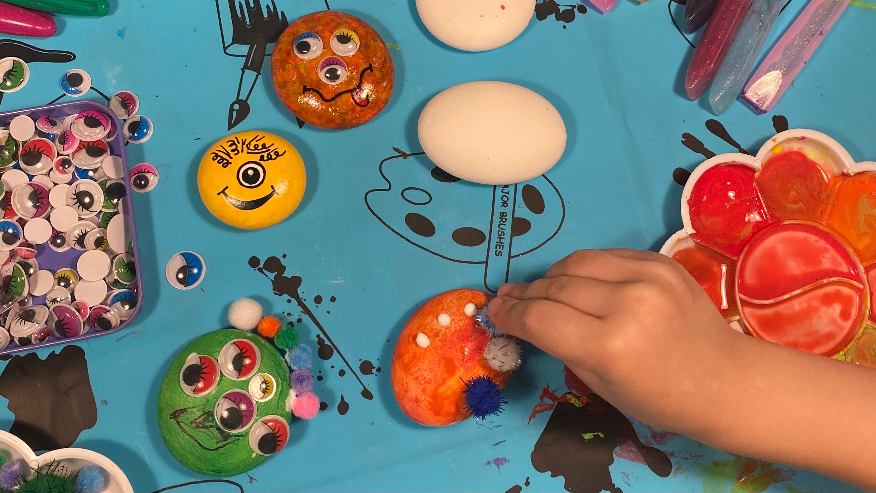 A hand glueing sparkly pom poms onto an orange painted pebble. Also in shot are some blank pebbles and other pebble monsters with craft materials including a tray of googley eyes spilling onto the table. 