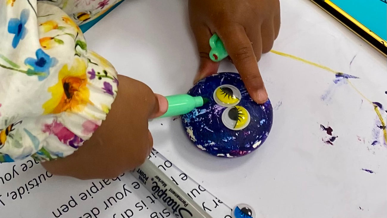 A child's hand drawing a mouth onto a purple painted pebble monster with yellow gooley eyes. Also in shot an instruction sheet for how to make pebble monsters.