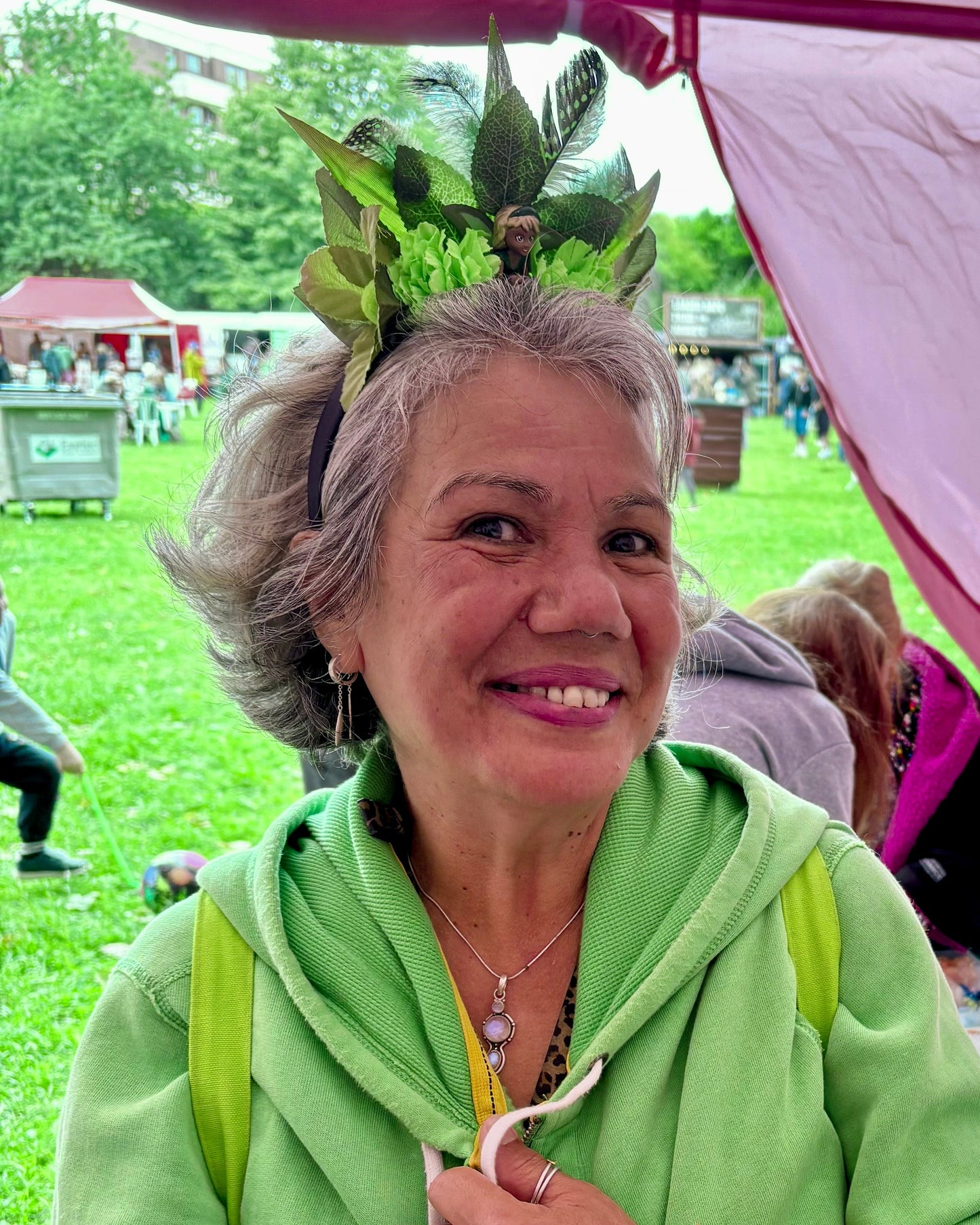 A smiling woman with gray hair wearing a green leafy headband and a light green zip-up hoodie stands under a pink market stall canopy, with a table of small crafts and beside her on a grassy outdoor market day.