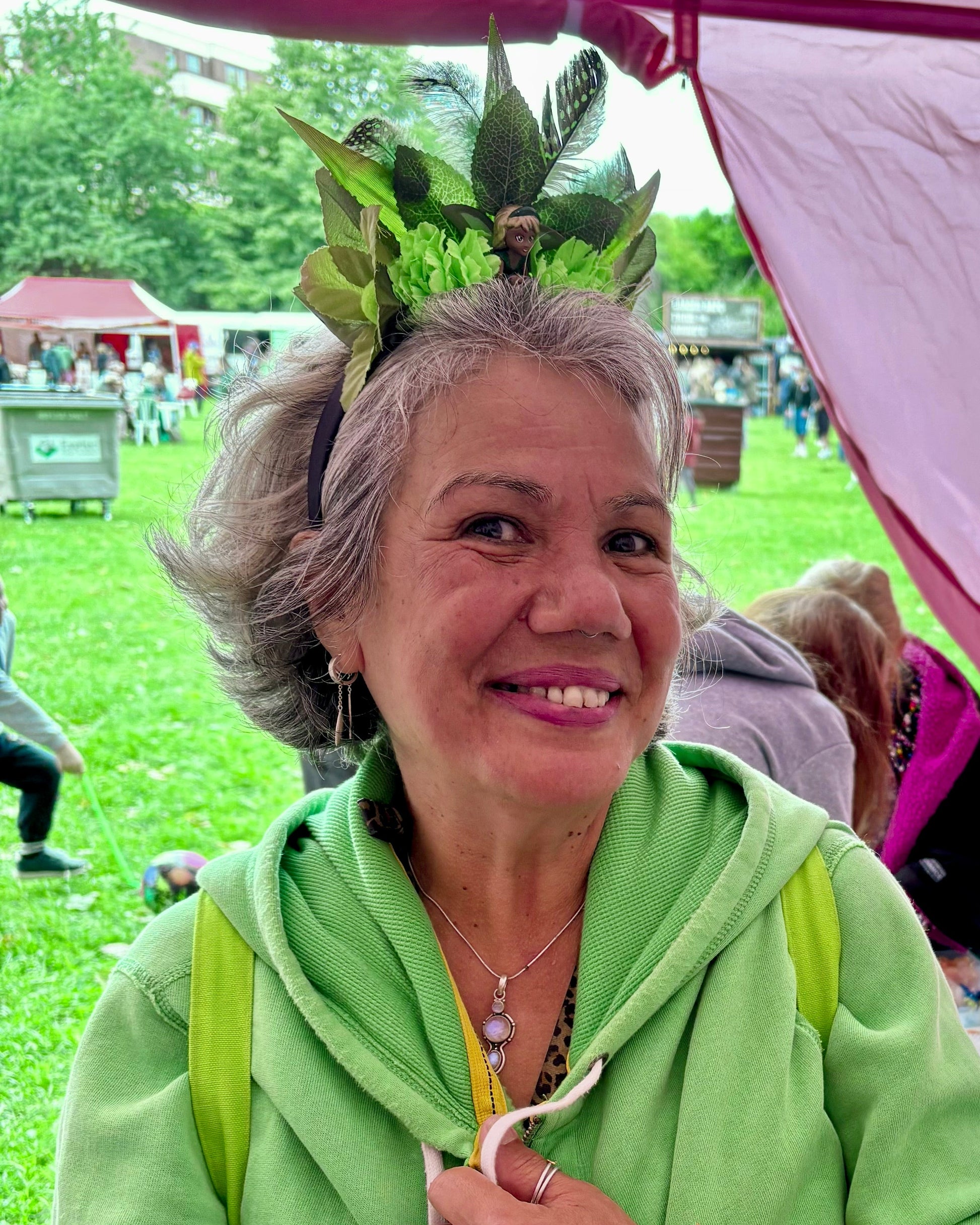 A smiling woman with gray hair wearing a green leafy headband and a light green zip-up hoodie stands under a pink market stall canopy, with a table of small crafts and beside her on a grassy outdoor market day.