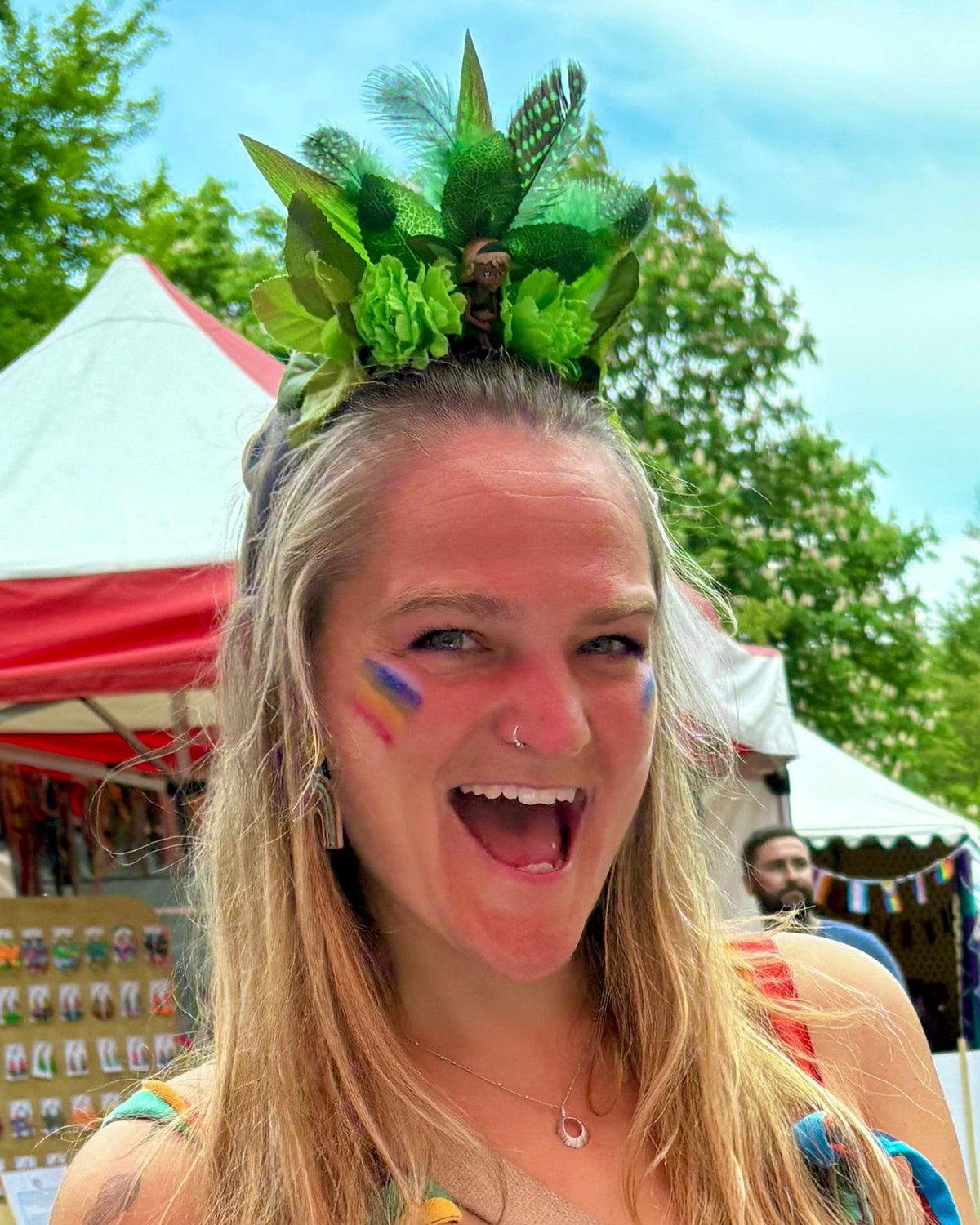 A woman at an outdoor festival wearing a green leaf and feather headpiece with a Pride flag painted on her cheek. Tents and trees are visible in the background.