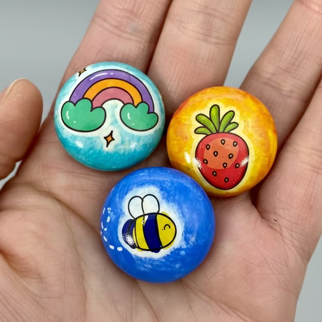 A hand holding 3 colourful mini pebbles with various designs on. A rainbow, a strawberry and a cartoon bee.
