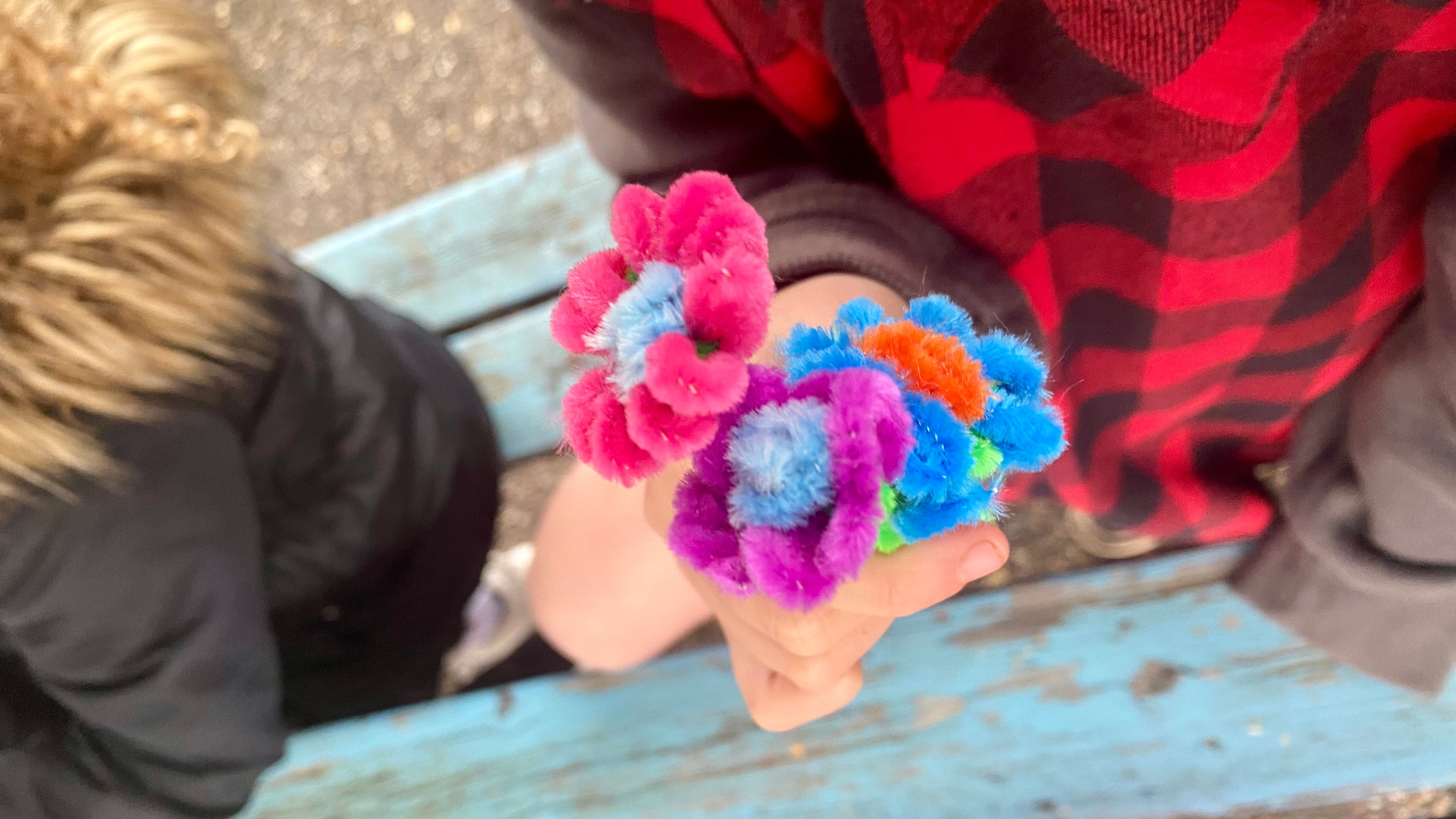 A child's hand holding 3 small flowers made from pipecleaners, the flowers are pink, purple, blue and orange with green stems.