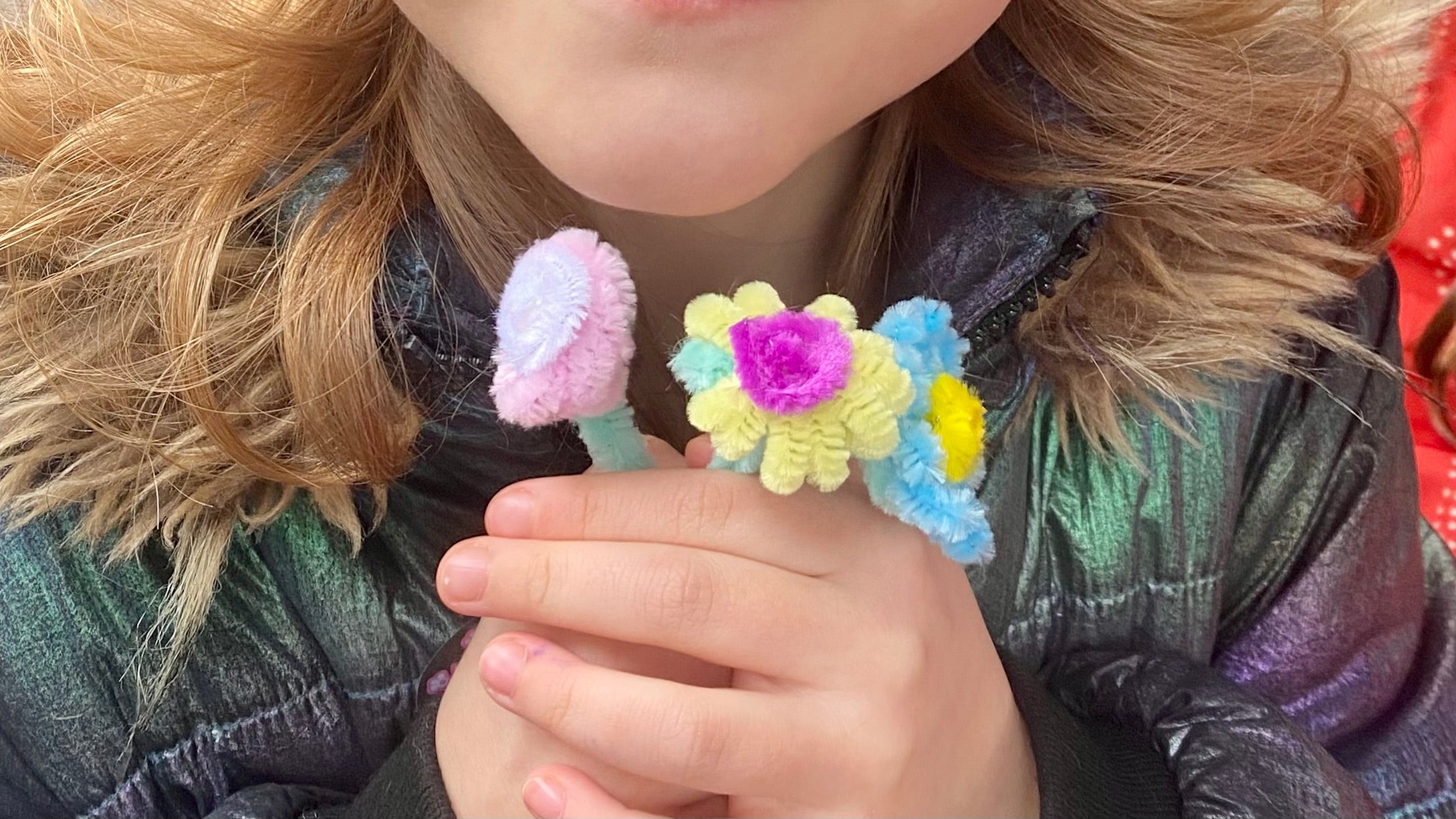 A child's hand holding 3 small flowers made from pipecleaners, each flower is made from 3 different coloured pipecleaners, one for the centre of the flower, on for the petals and one for the stem.