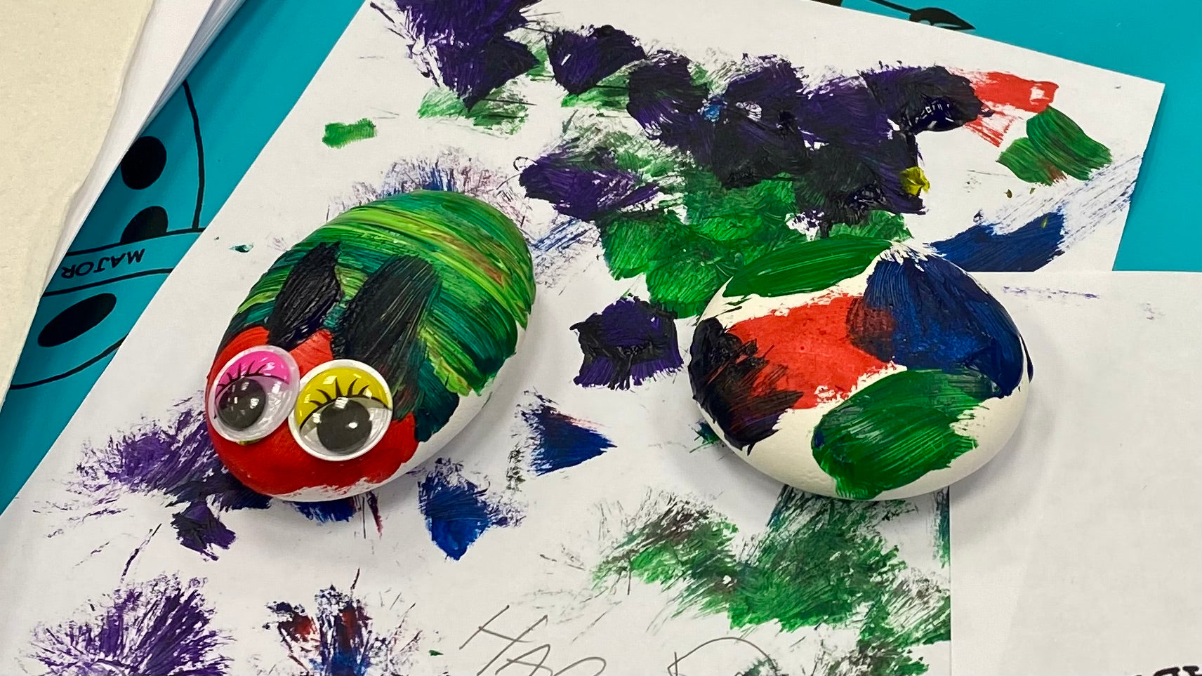 2 painted pebbles drying on a piece of paper covered in paint marks. One of the pebbles is painted to look like the Very Hungry Caterpillar and has brightly coloured googly eyes stuck on. The other pebble is covered in bright paint brush strokes.