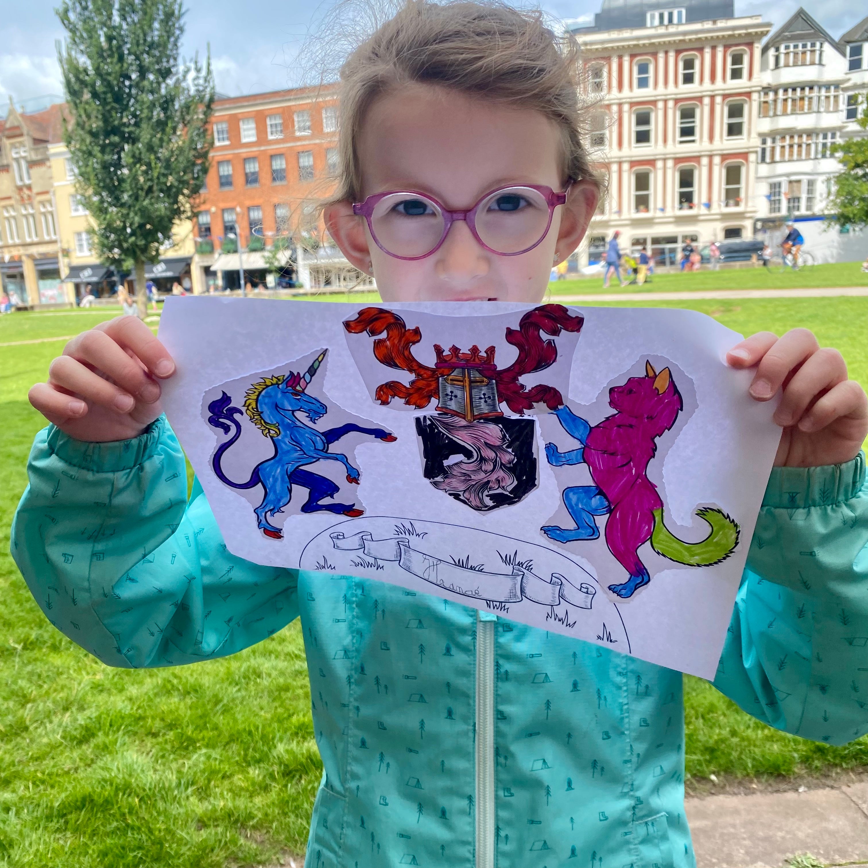 A girl holding up a sheet of paper with a custom crest on. The crest has a unicorn, a cat and other motifs and has been coloured in using bright colours