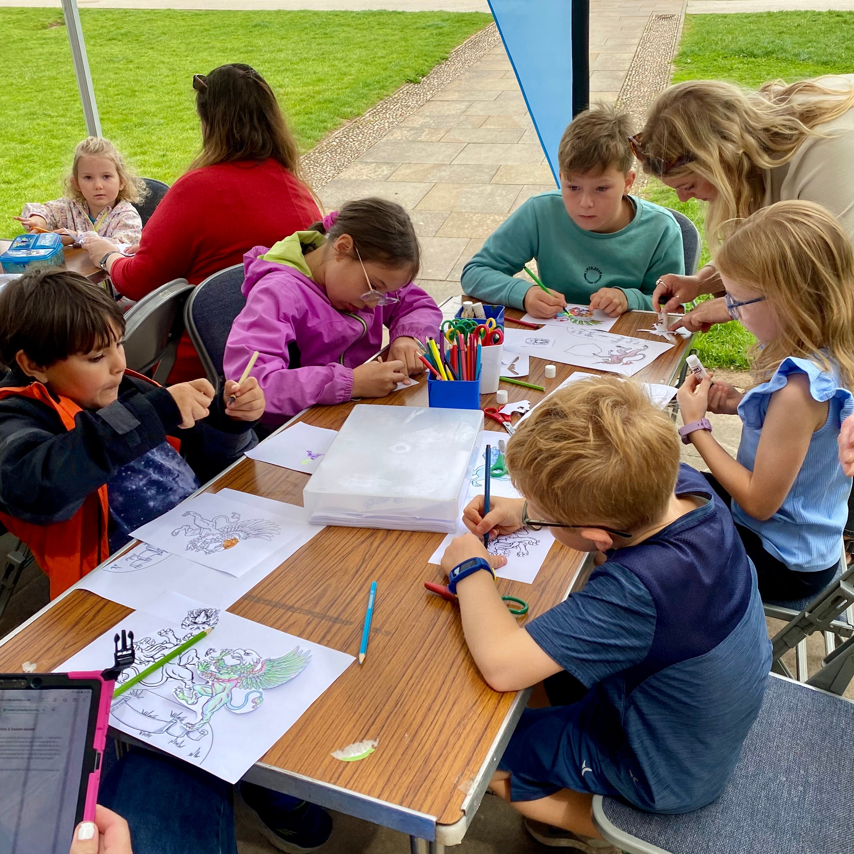 An outdoor workshop where children are sat around tables under a gazebo creating their own crests by cutting, sticking and colouring.
