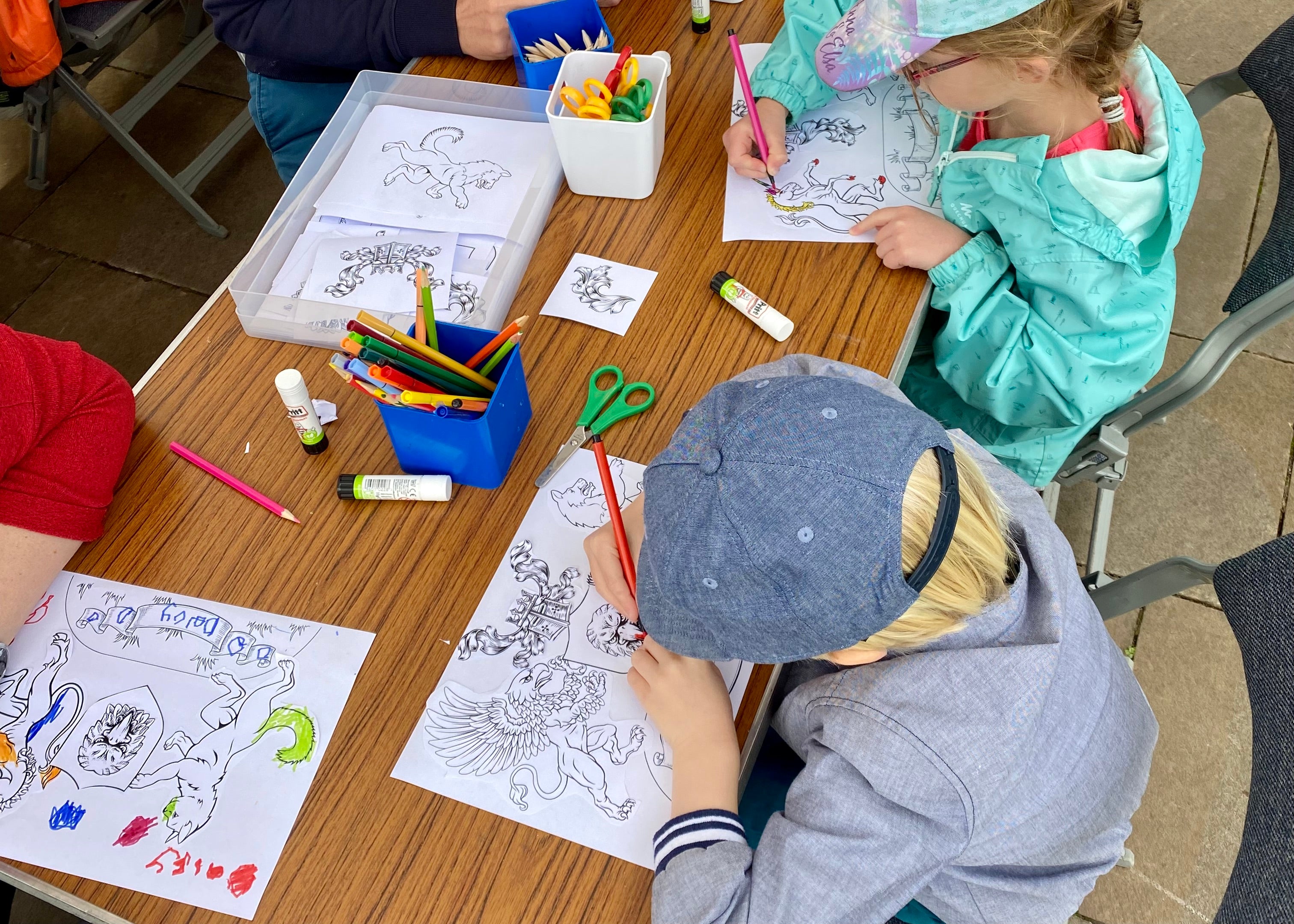 An outdoor workshop where children are sat around tables under a gazebo creating their own crests by cutting, sticking and colouring.