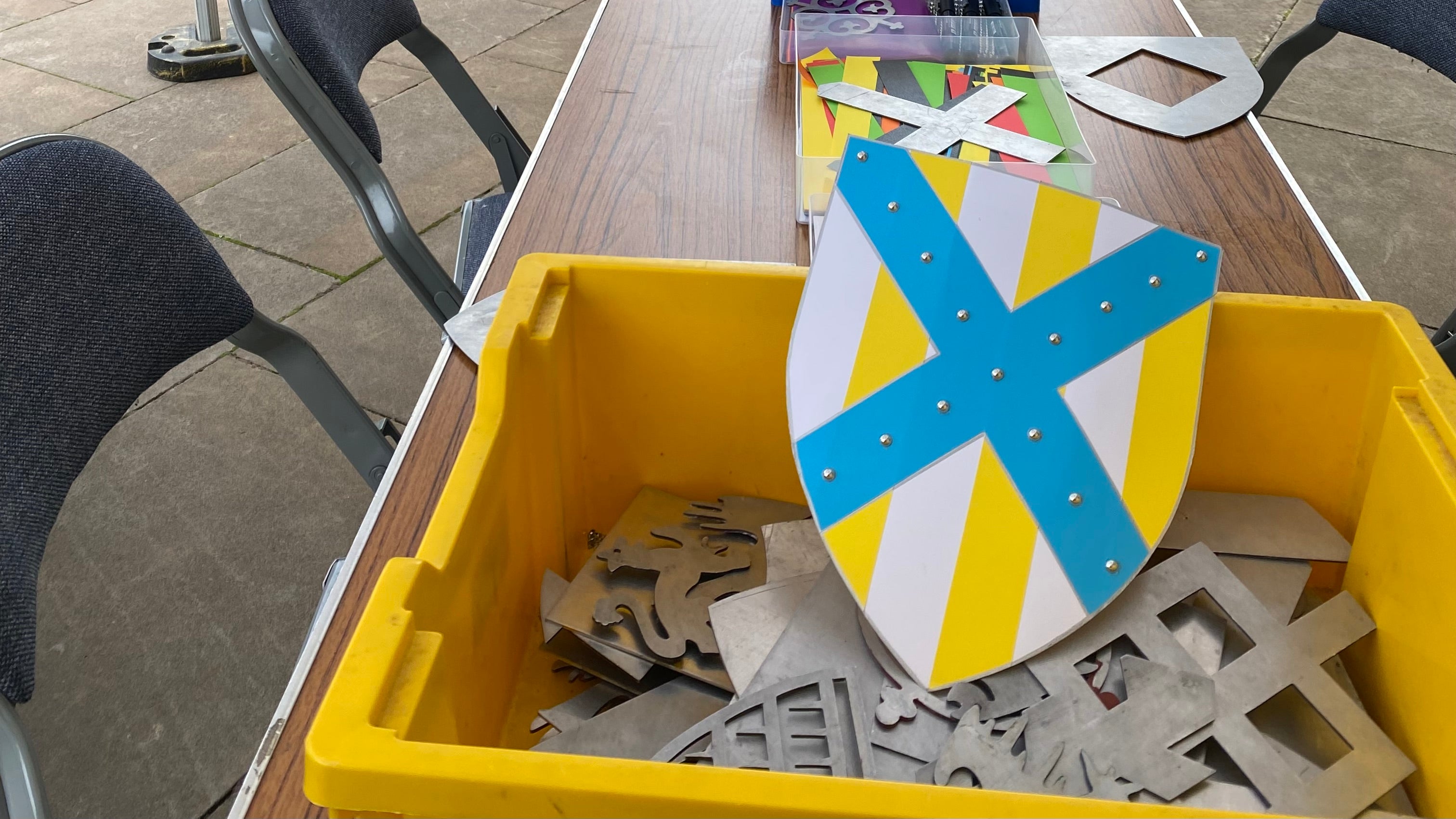 A cardboard shield in a box of metal templates. The shield has a blue diagonal cross over the top of yellow and white stripes, there are small silver stud stickers in the cross and silver outlines to the sections. In the background is a table with more craft materials on