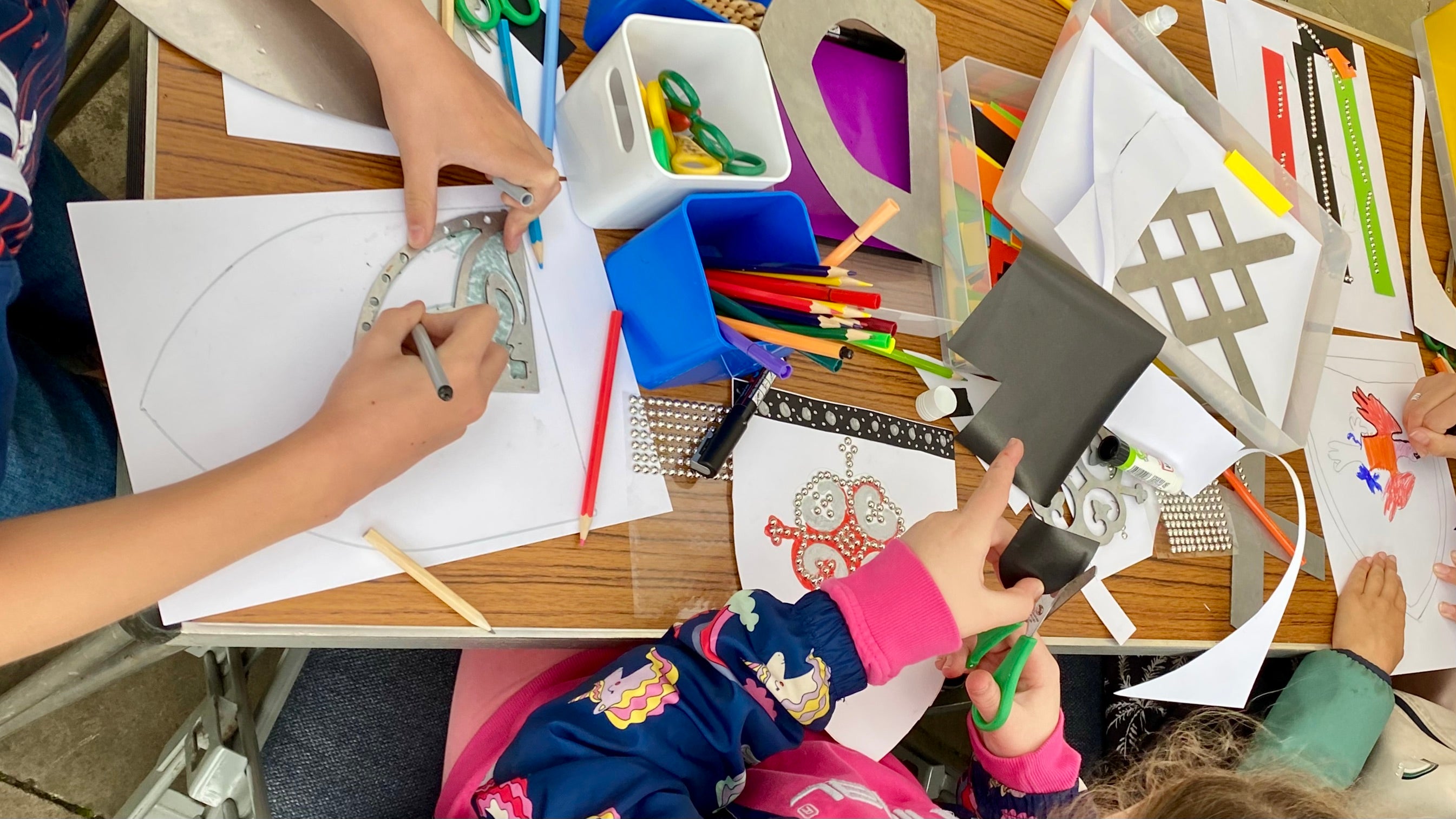 A workshop table with children crafting cardboard shields. There are craft materials strewn across the table including metal templates, colouring pencils, scissors and sticky silver studs. In shot are three children cutting and colouring