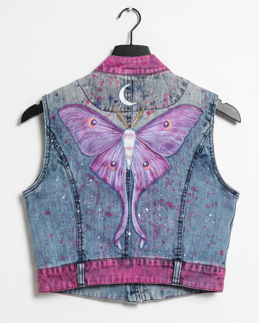 A sleeveless denim vest with a large purple and pink moth painted on the back, the vest has pink painted along the edges and a small crescent moon at the top.