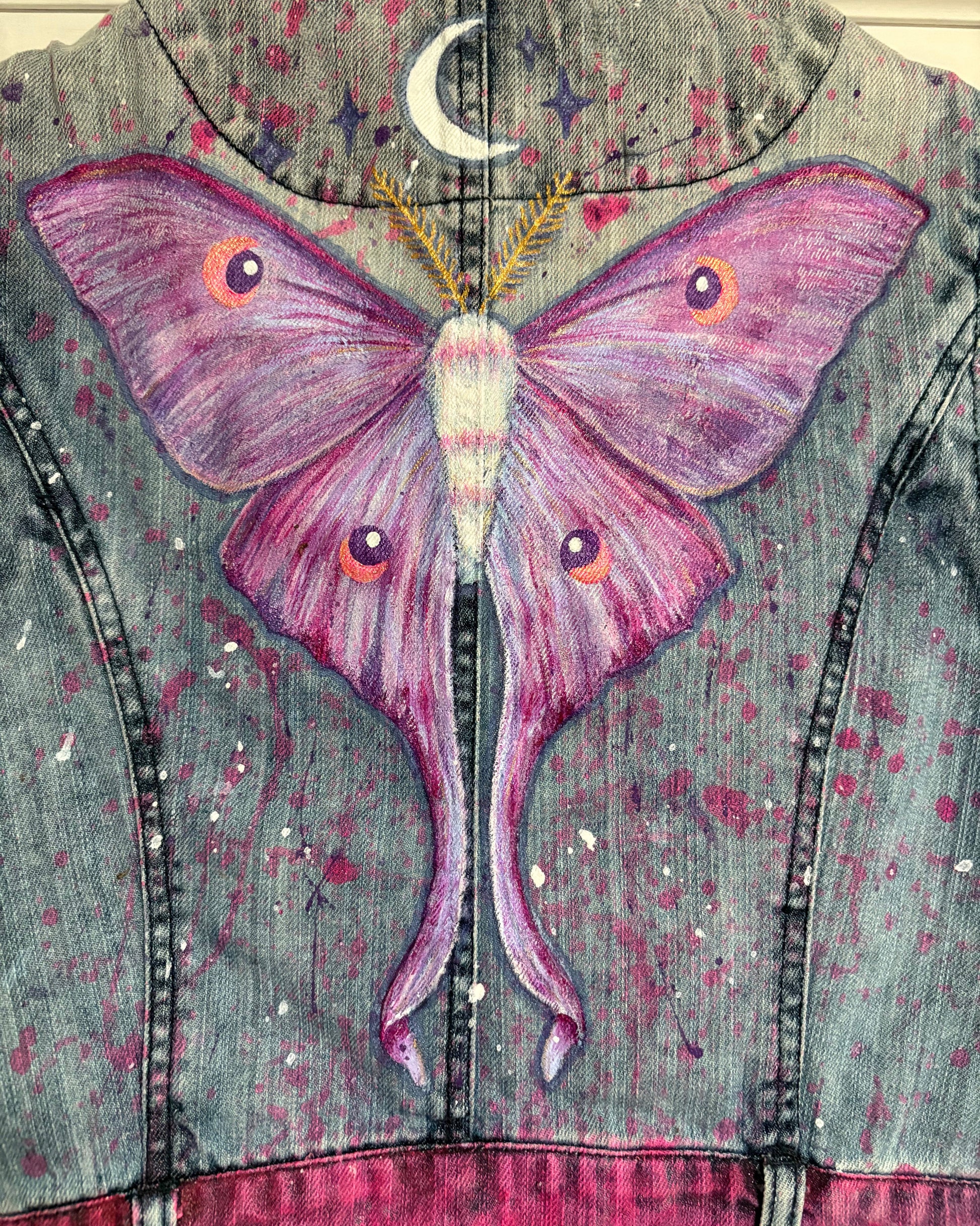 A close-up of the back of a denim jacket featuring a large painted purple and pink moon moth, with a crescent moon and stars on a distressed gray-blue denim background with pink paint splatter details.