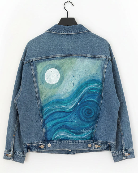 Denim jacket with a painted design of a moon and swirling ocean wave patterns on a white surface.