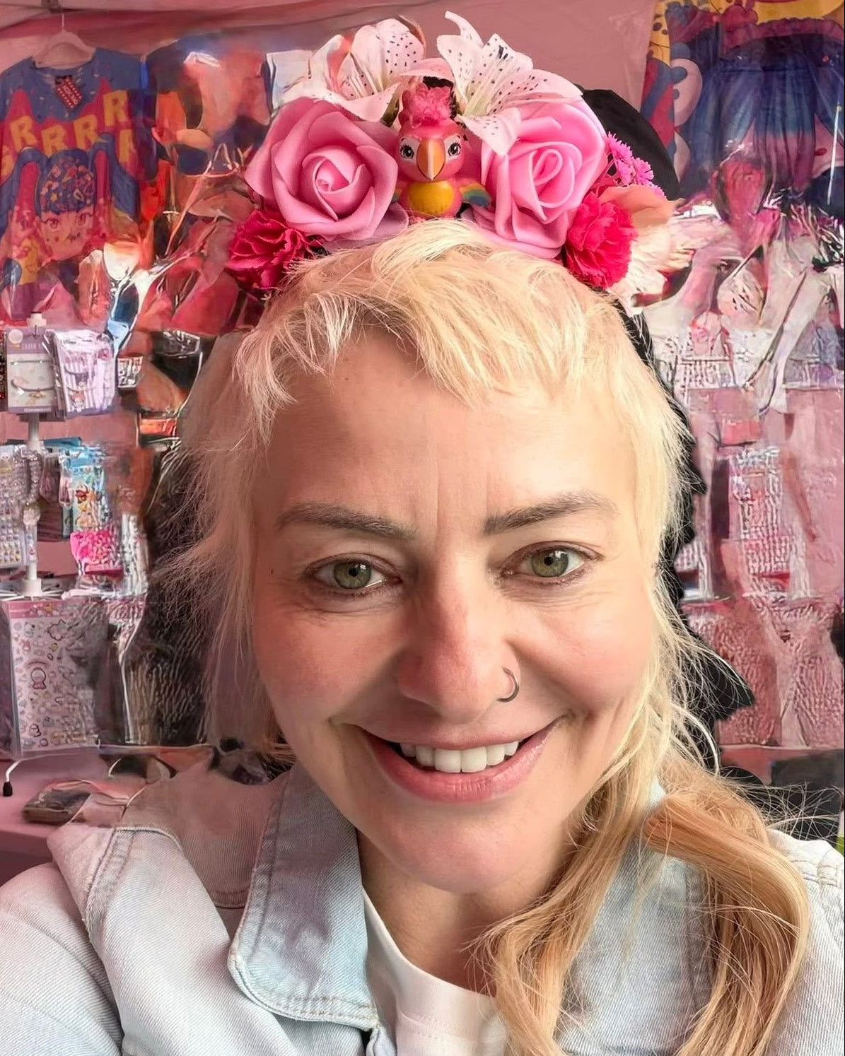 A smiling blonde woman takes a selfie wearing a large pink floral crown with roses and a small doll accessory, standing at a market stall filled with clothing and packaged items.