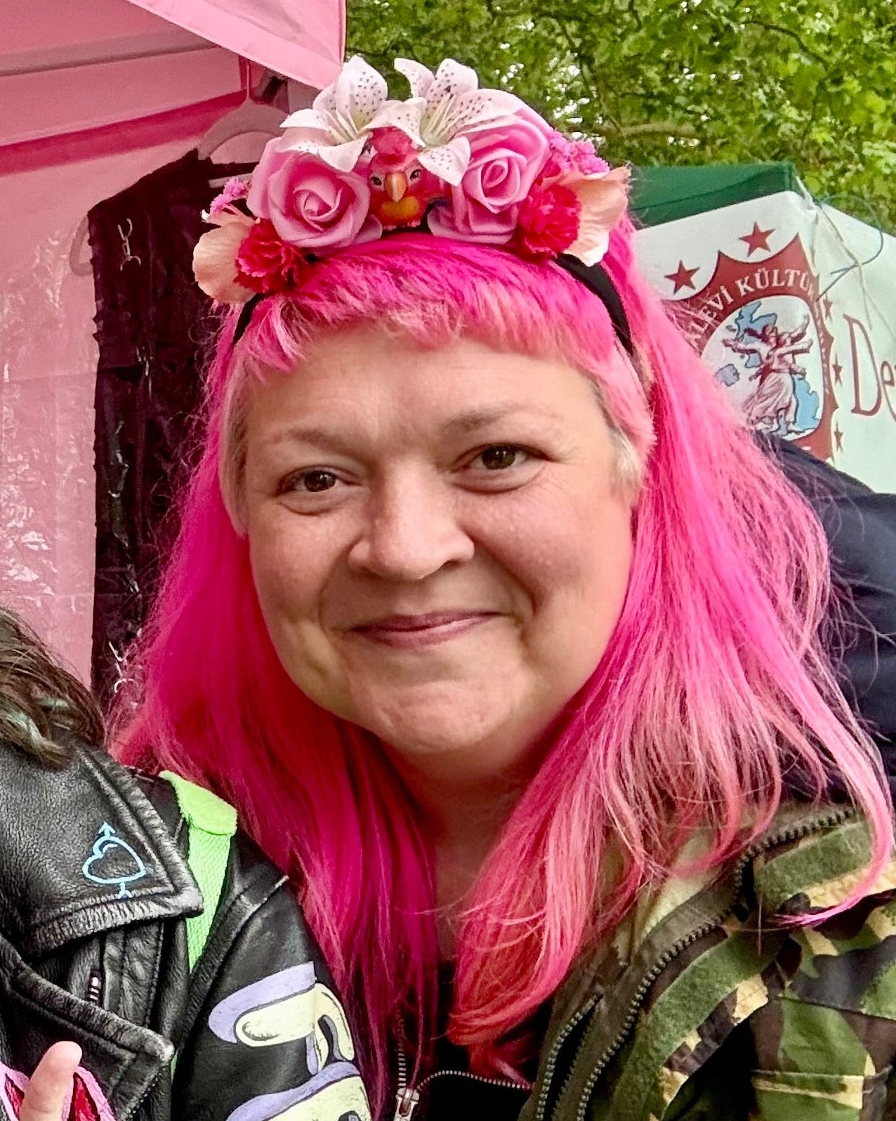 A smiling woman with bright pink hair wearing a pink floral headband photographed outdoors at a festival with tents and greenery in the background.