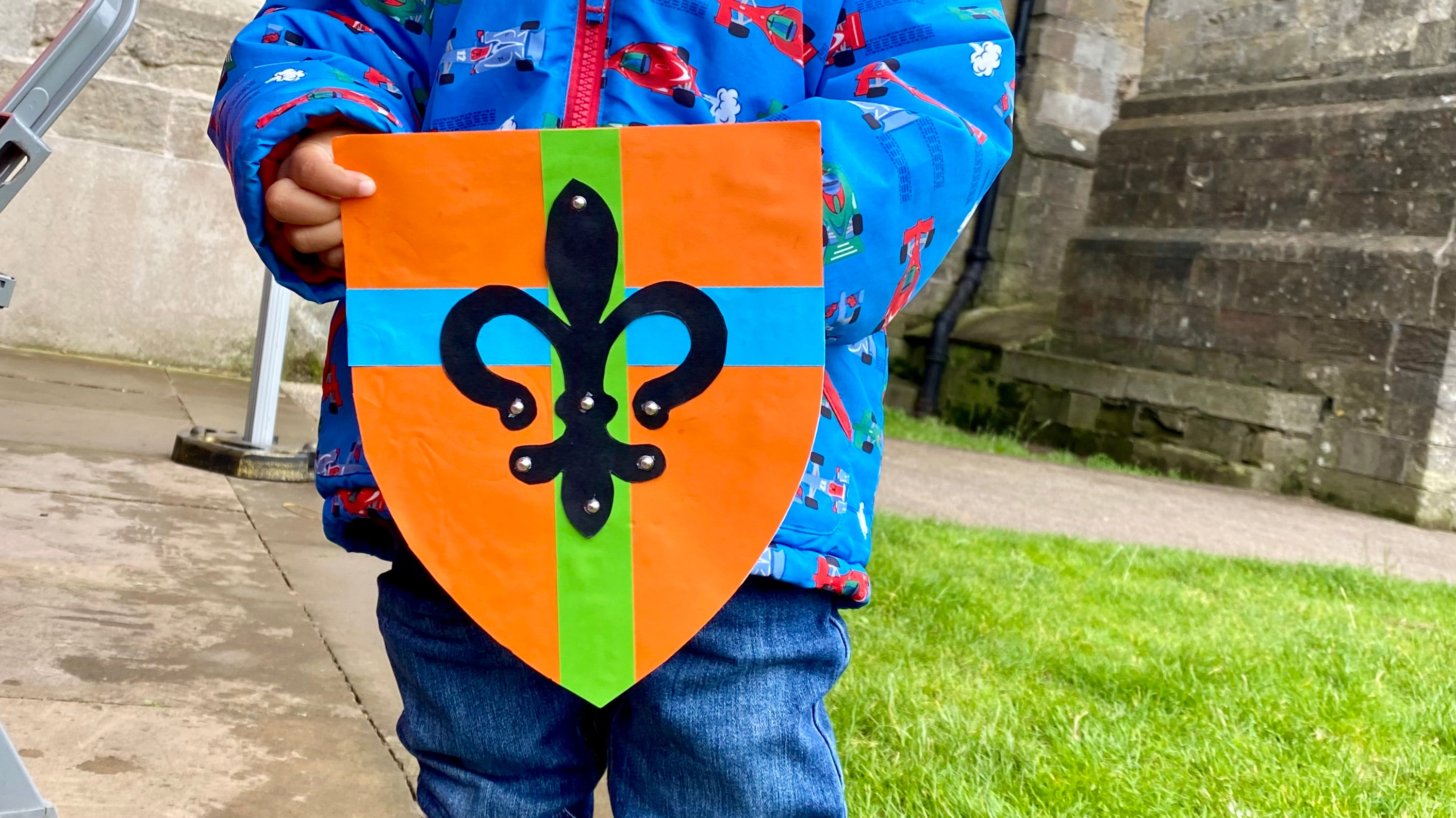 A cardboard shield being held by a child. The shield is orange with a green and blue stripe and a black filigree shape in the middle with small silver stud stickers on. In the background is the cathedral