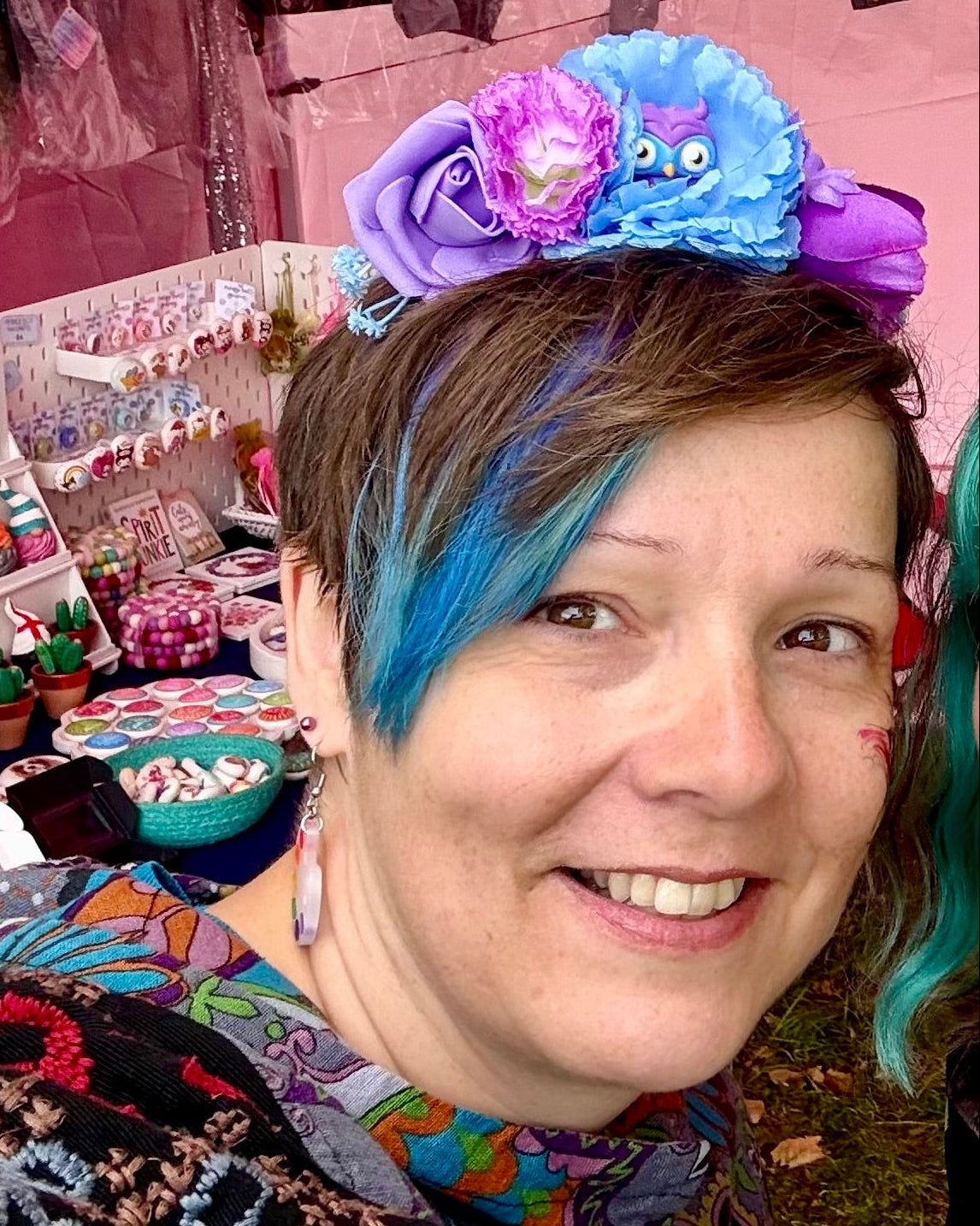 A smiling woman with short brown hair and bright blue side streaks wears a colorful handmade flower headband with purple and blue flowers, and a small owl figure. She is photographed at a craft fair with various colourful items in the background.