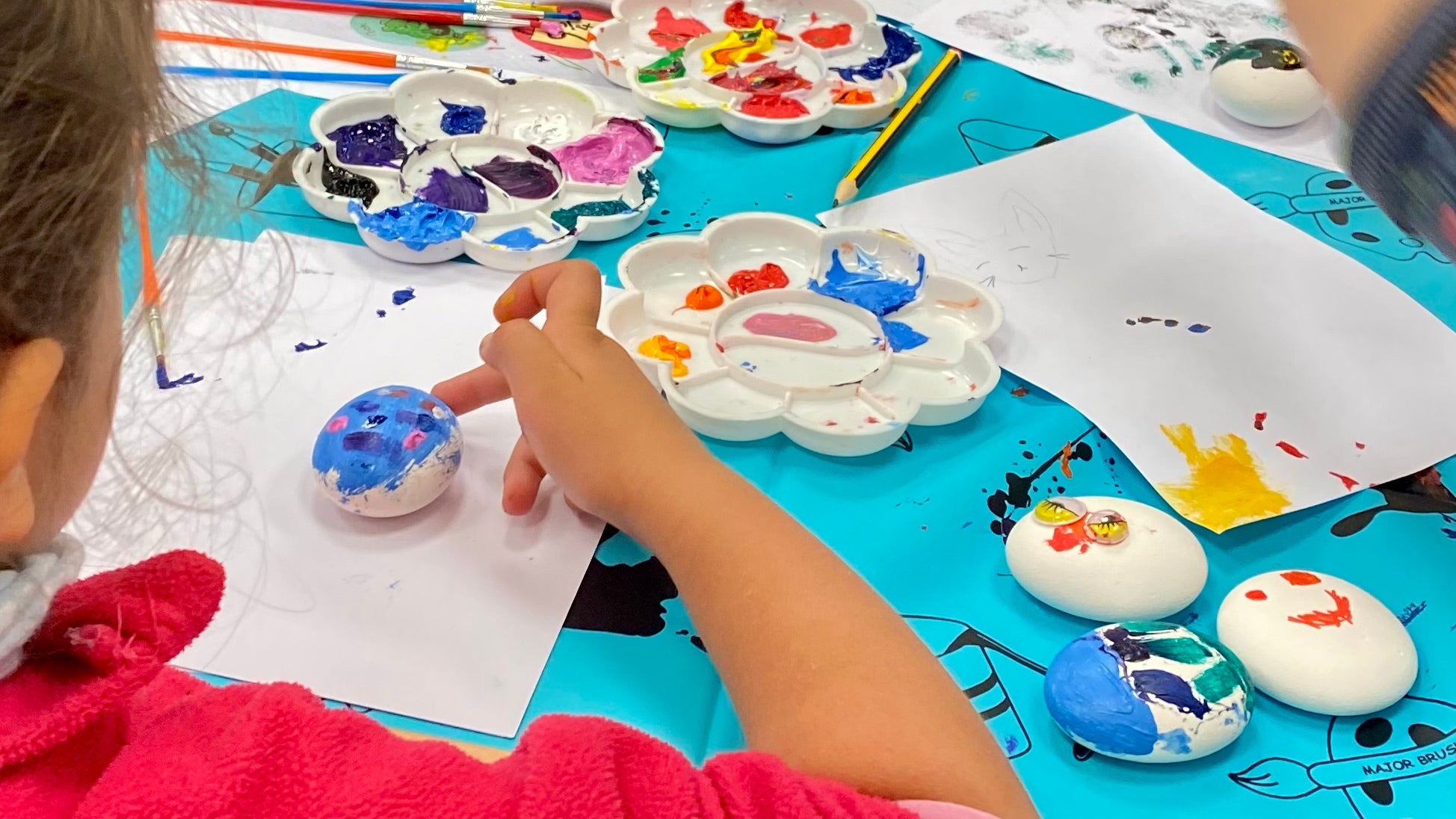 Table covered in painted pebbles, paint palettes, brushes, pencils and pieces of paper. 2 children are actively painting their pebbles.