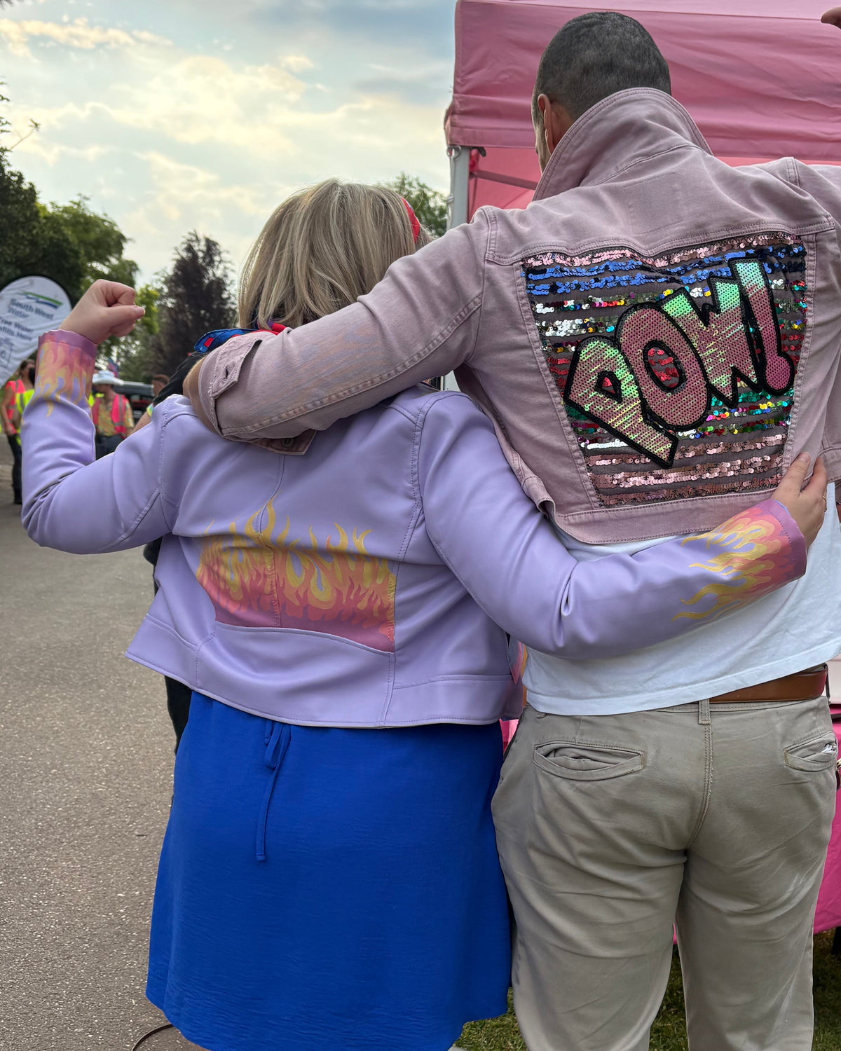 Two people standing outdoors with their backs to the camera, one wears a lavender cropped PU jackets painted with pastel flames on the back and sleeves. A pink event tent and tables are visible in the background.