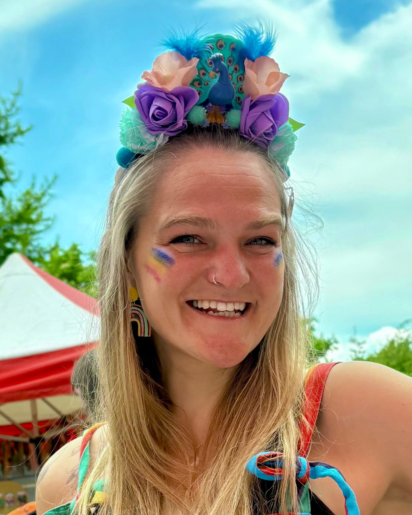 A smiling woman outdoors wearing a colorful floral headband with blue, purple, and pink flowers with a toy peacock. Trees, a red-and-white tent, and blue sky are visible in the background.