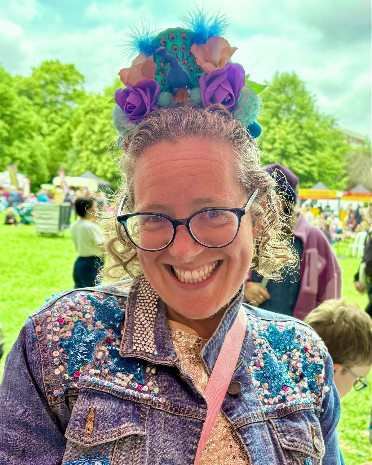 A smiling woman at an outdoor festival wearing a colorful floral headpiece featuring a toy peacock, round glasses, a blue denim jacket with pink and blue floral embroidery, and a beige knit top. She is standing under a pink canopy with a grassy fairground in the background.