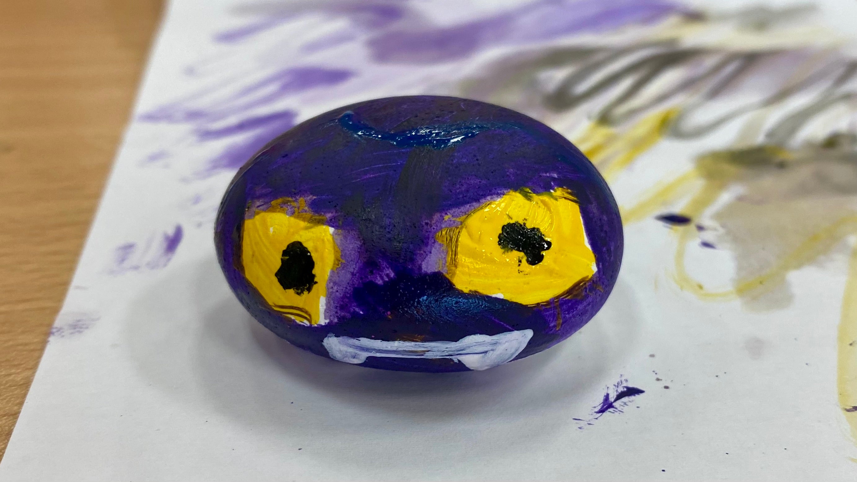 A pebble painted purple with menacing yellow eyes and a white mouth with fangs. The pebble sits on a piece of paper with paint and pen marks on.