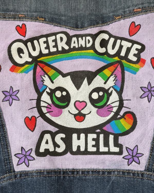 Close up view of a pastel purple panel on a hand-painted upcycled denim jacket featuring a rainbow cat, hearts, flowers, and the words “Queer and Cute as Hell”