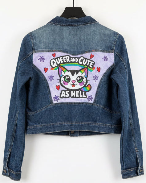 Back view of a hand-painted upcycled denim jacket with a pastel purple panel featuring a rainbow cat, hearts, flowers, and the words “Queer and Cute as Hell”