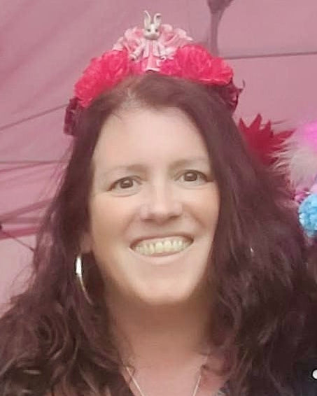 A women posing with friends under a pink canopy tent, wearing a colorful red floral crown headpiece with rabbit toy and vibrant, patterned dress.