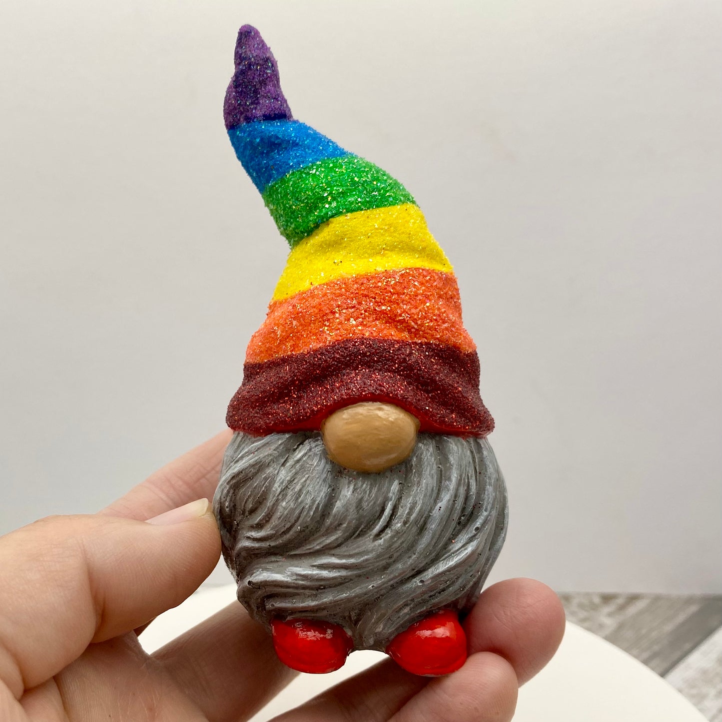 Hand holding a Small hand painted gonk statue with a Glittery Rainbow hat, grey beard and red shoes. Front view.