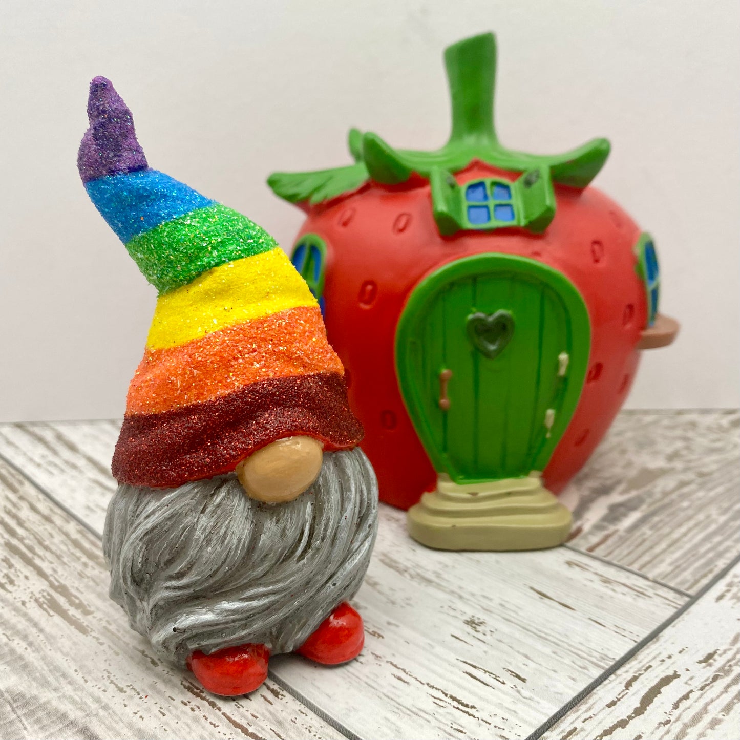 A small hand painted gonk statue with a Glittery Rainbow hat, grey beard and red shoes. Behind him stands a strawberry fairy house.