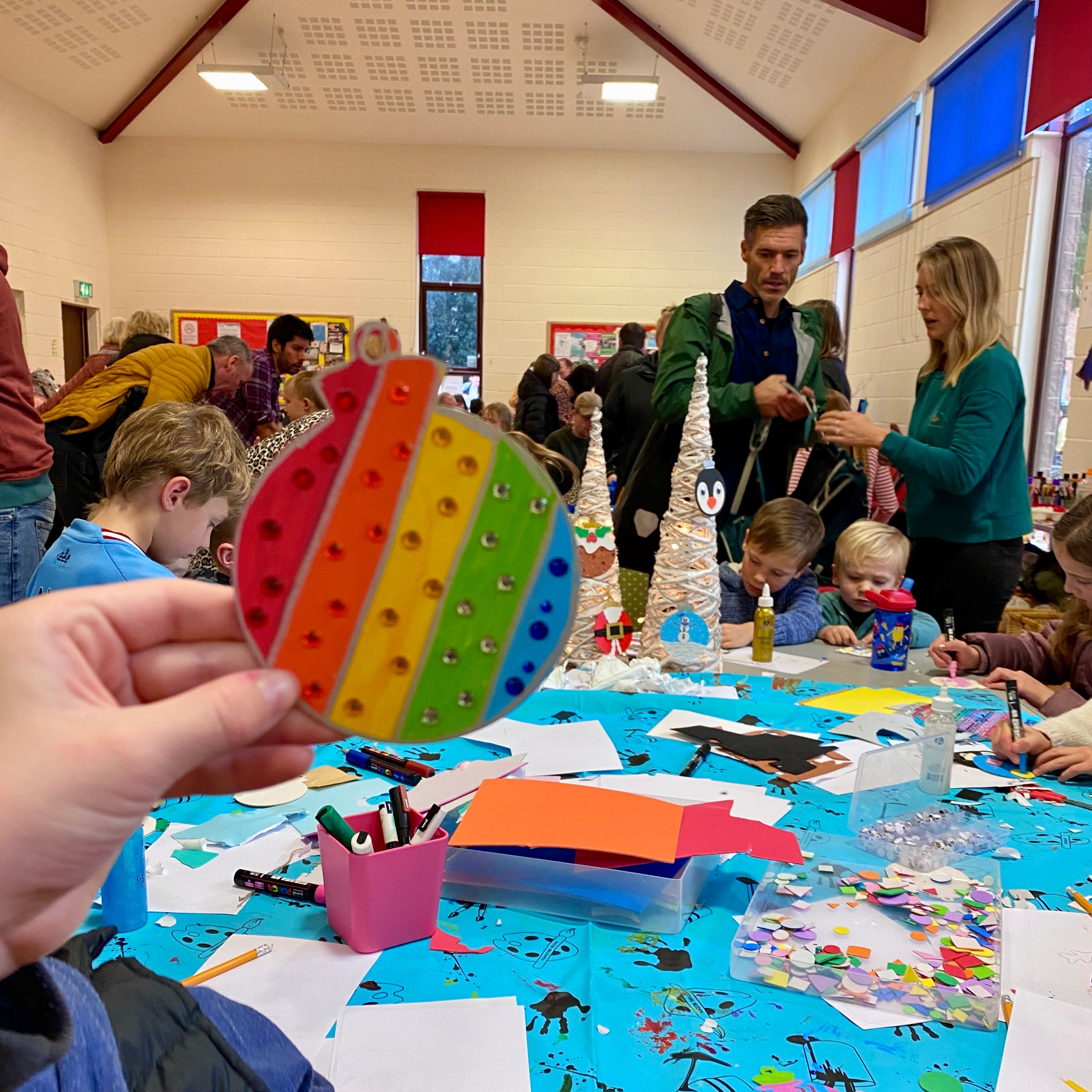 A Rainbow painted bauble with sparkly gems held up in front of a crafting table where a workshop is in full swing with children sat around a table creating their own Christmas baubles