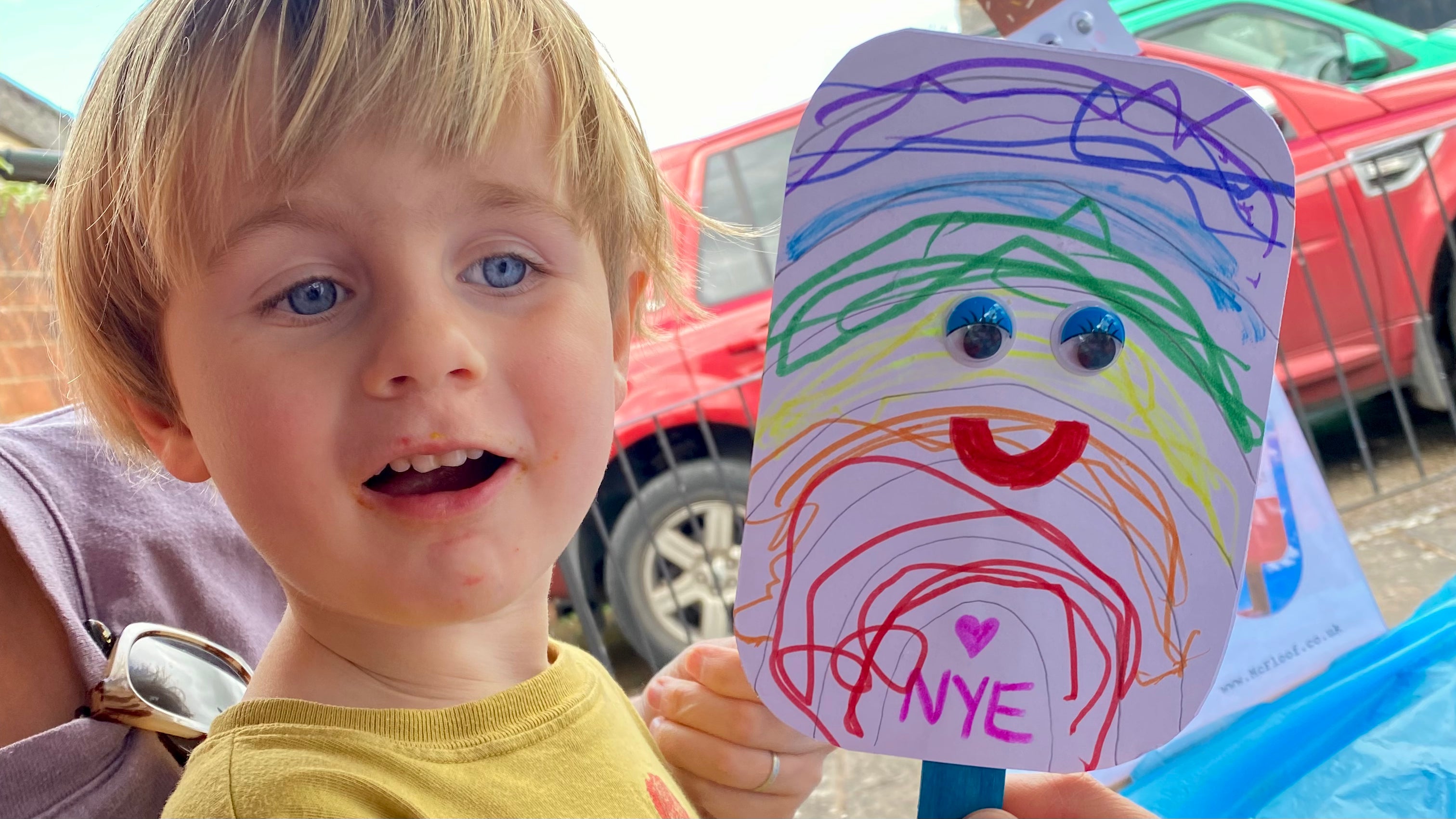 Child proudly holding their ice lolly card creation. The card is on a blue wooden stick and has a rainbow drawn on with a smiley face with googley eyes.