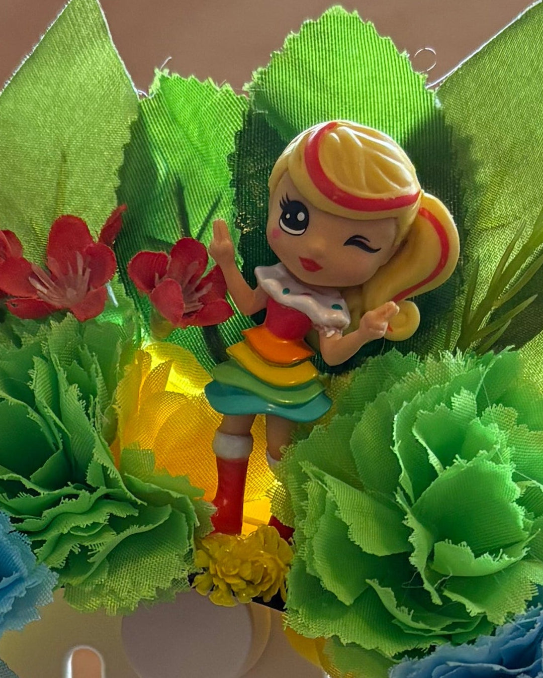 A close up of a colourful floral headband featuring green leaves, green and red flowers, and a girl figurine wearing a rainbow dress and red boots.