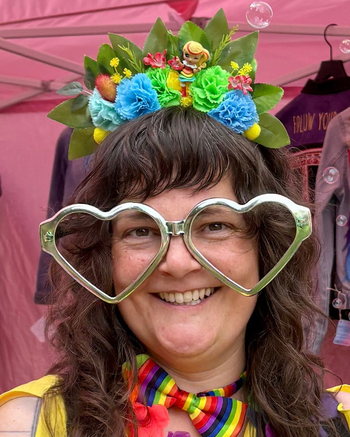 A smiling woman at a festival booth wearing a rainbow themed flower crown and large heart-shaped glasses, with a pink canopy and colourful clothing behind her.