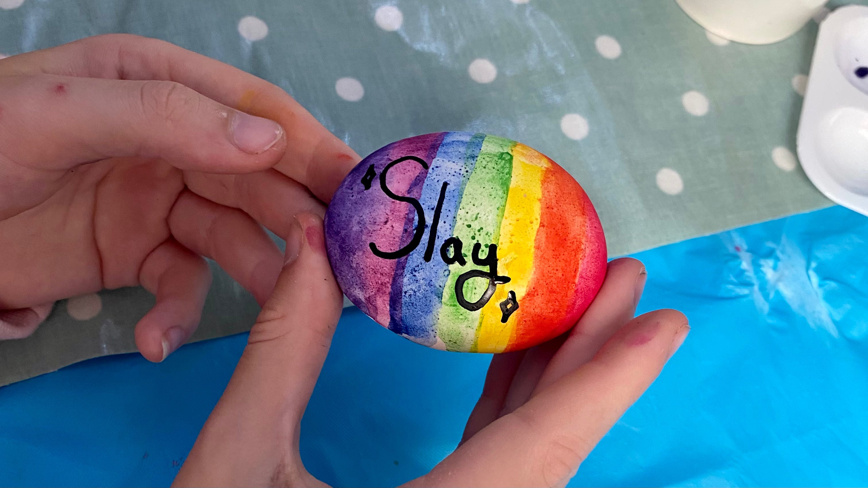 Hands holding a rainbow painted pebble with the word Slay written on in black
