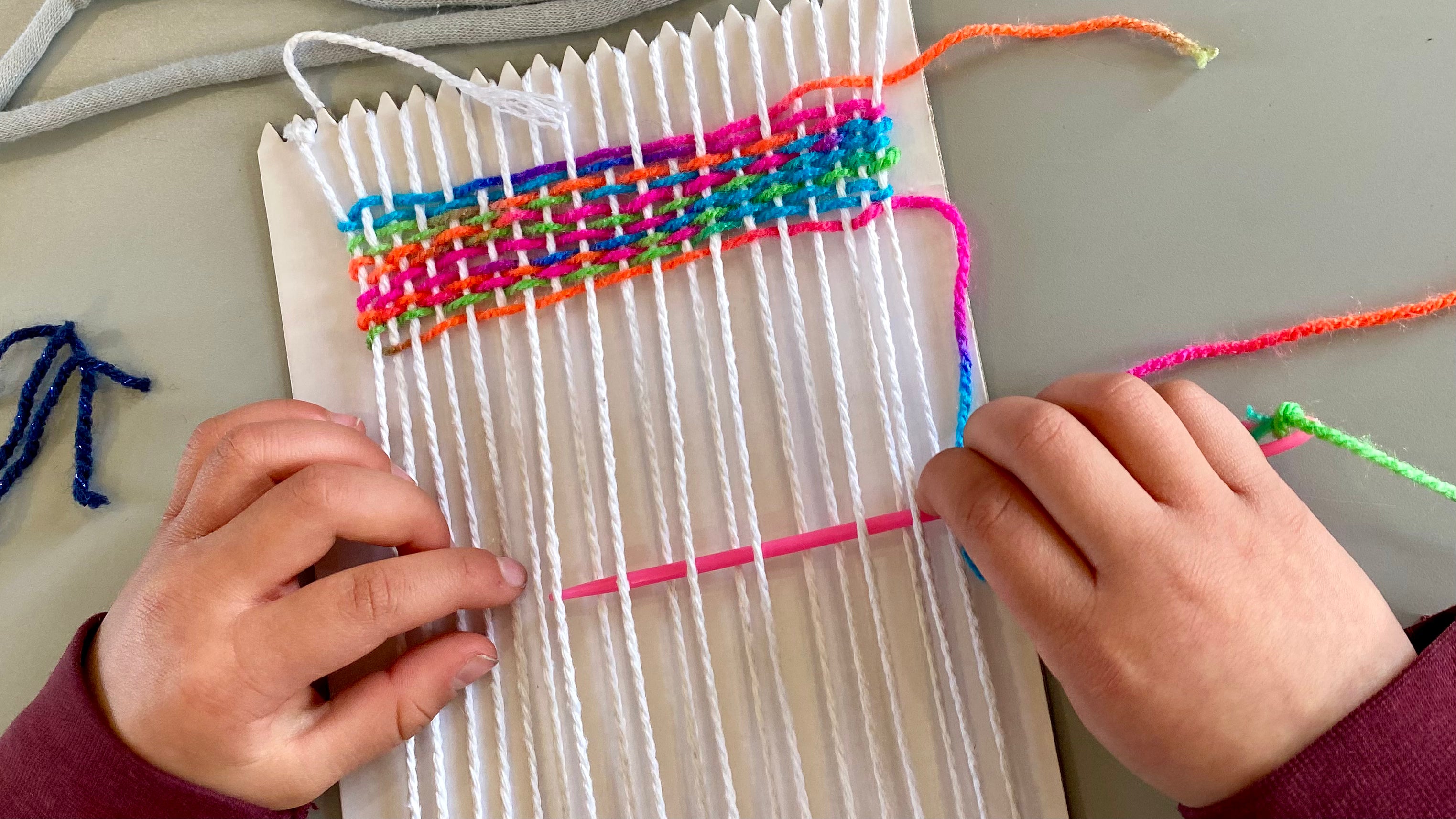 A pair of child's hands holding a large pink needle threading bright rainbow wool in and out of thread on a cardboard weaving loom
