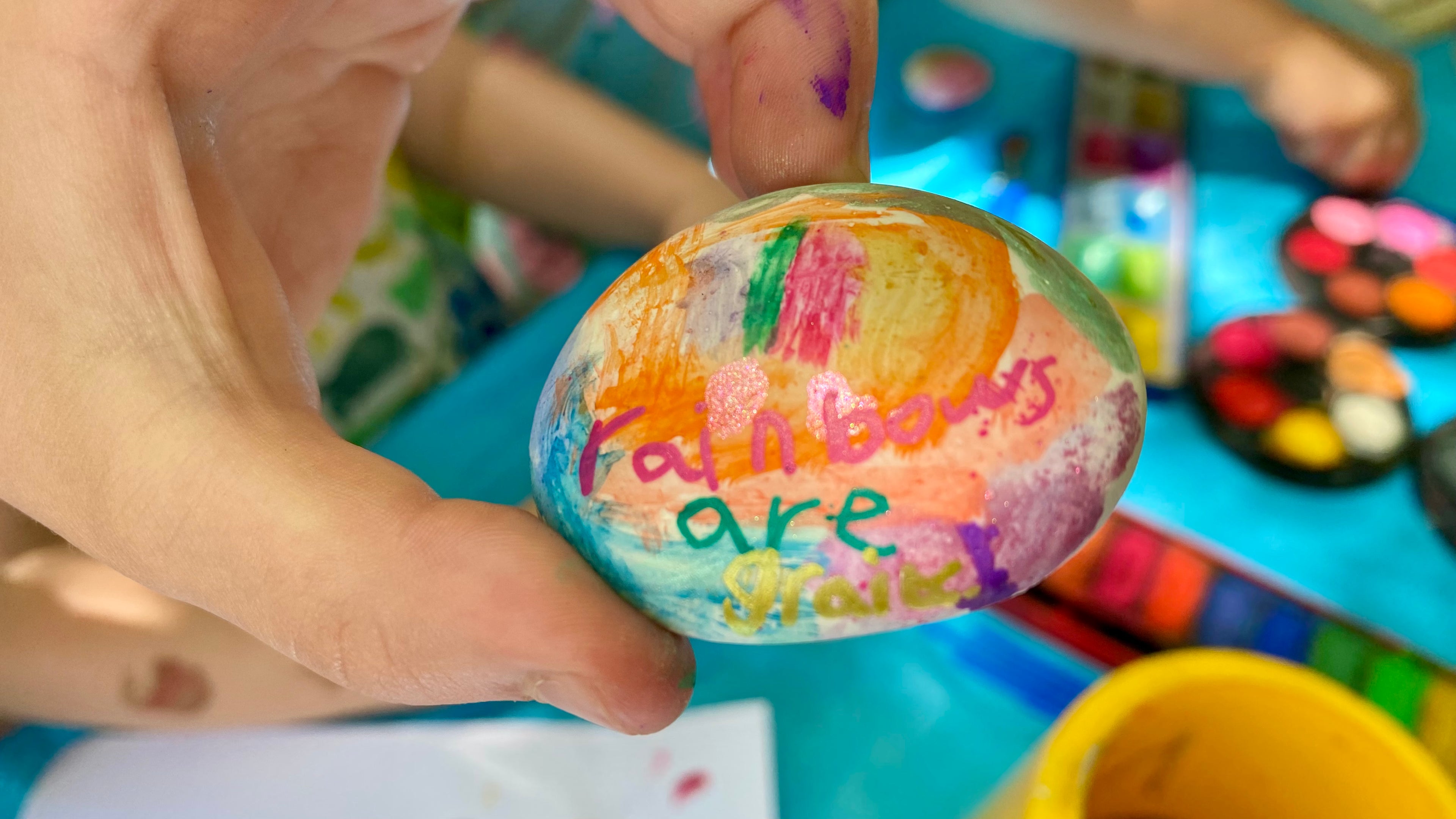 Hand holding a brightly painted pebble with the words Rainbows are Great written on. In the background there's a table with pallettes of watercolour paints