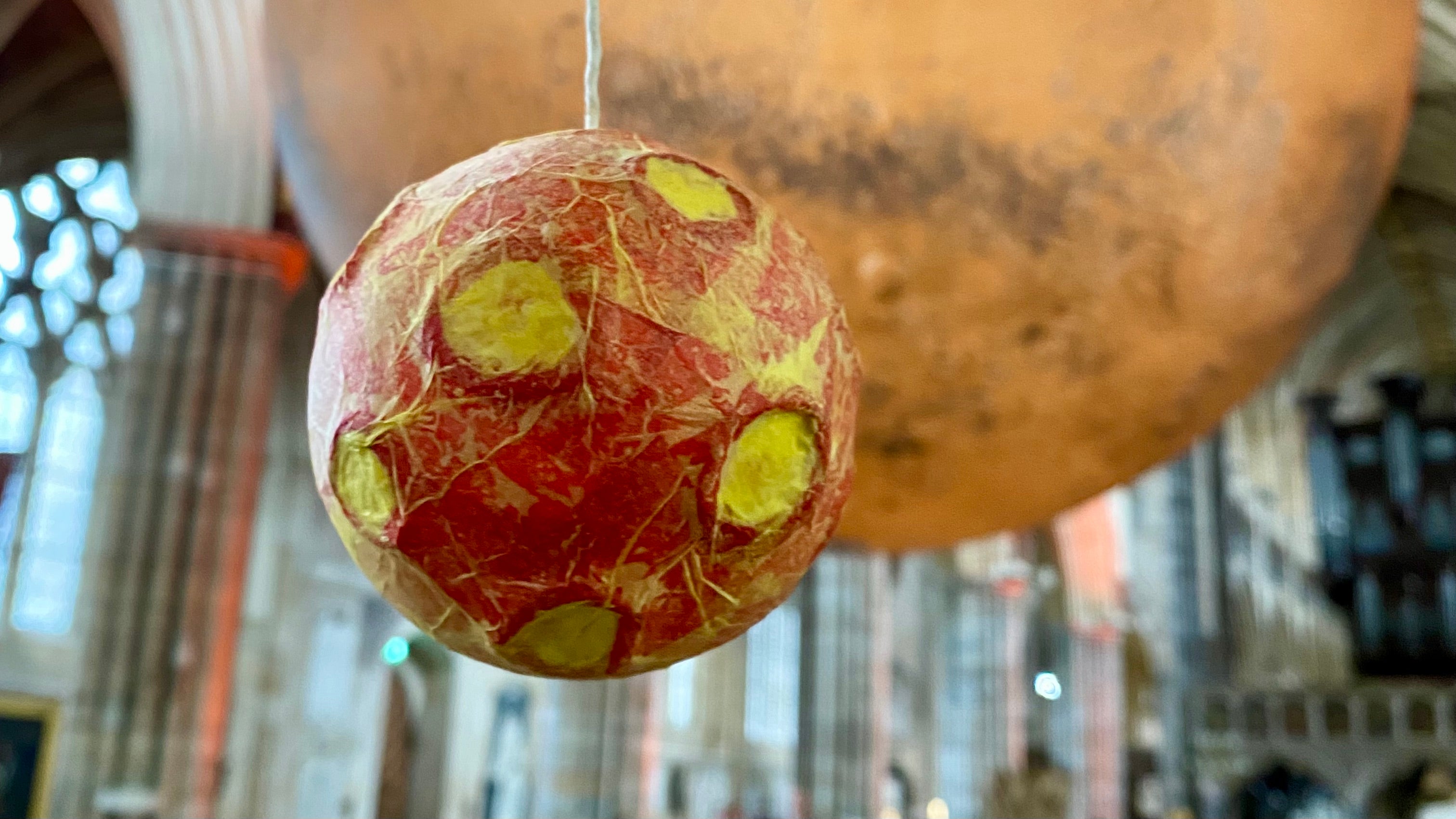 A handmade paper mache red planet with yellow craters hanging from a string in a cathedral with a large moon installation behind.