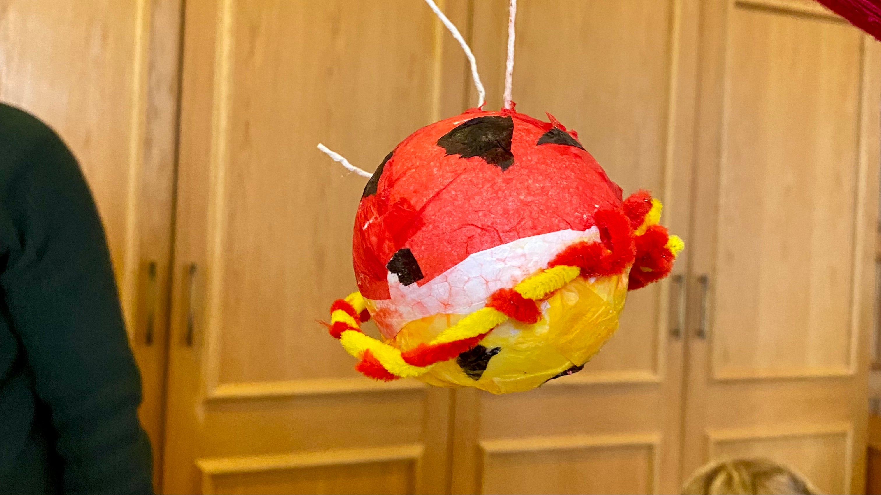 A paper mache planet being held up in front of some doors. The planet is made from paper mache and is red and yellow with black patches. There is a ring around the planet made from a red and yellow pipe cleaner twisted together.