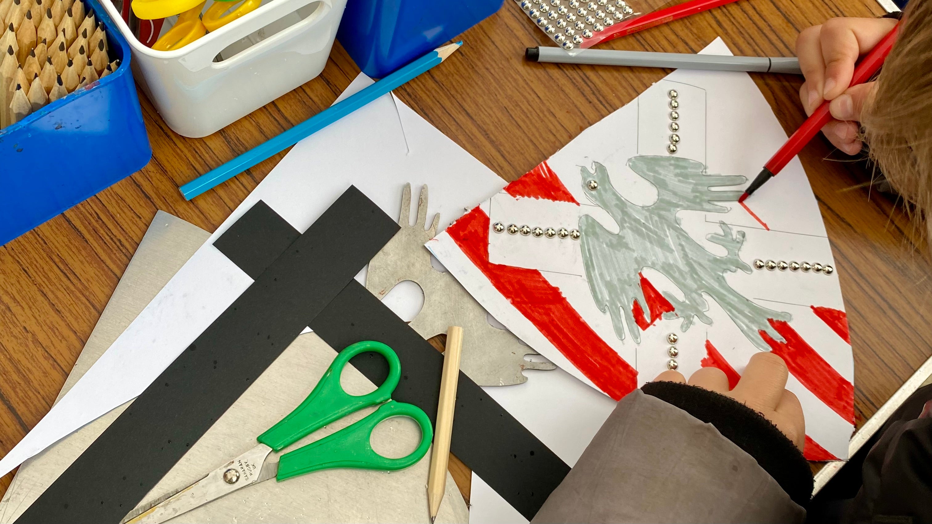 A cardboard shield in the process of being coloured in. The shield is white with vertical red stripes and a white diagonal cross with a silver dragon in the centre of the shield. There are small silver stud stickers in the cross. The table has craft materials on