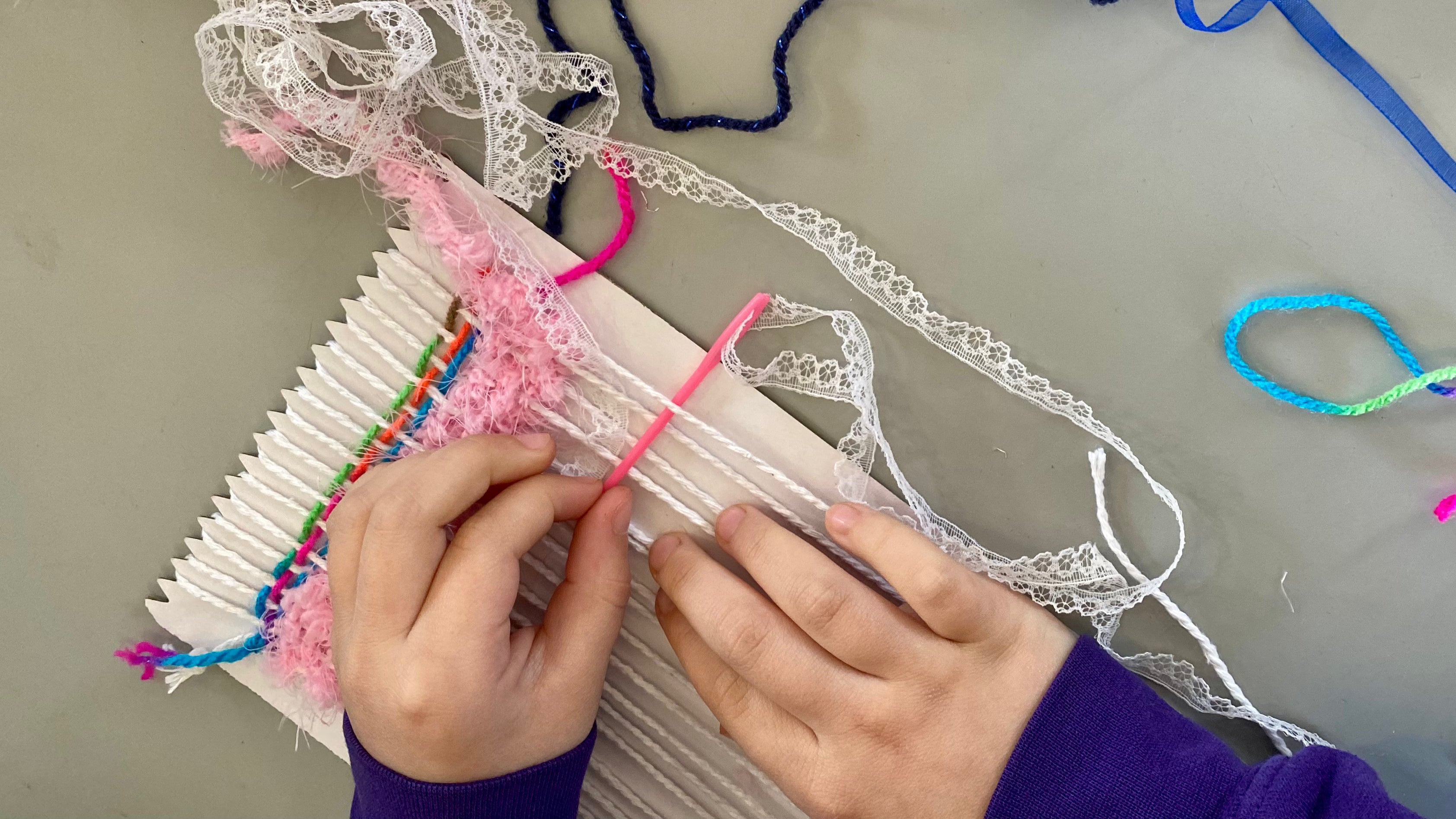 A pair of child's hands holding a large pink needle threading lace trim in and out of thread on a cardboard weaving loom