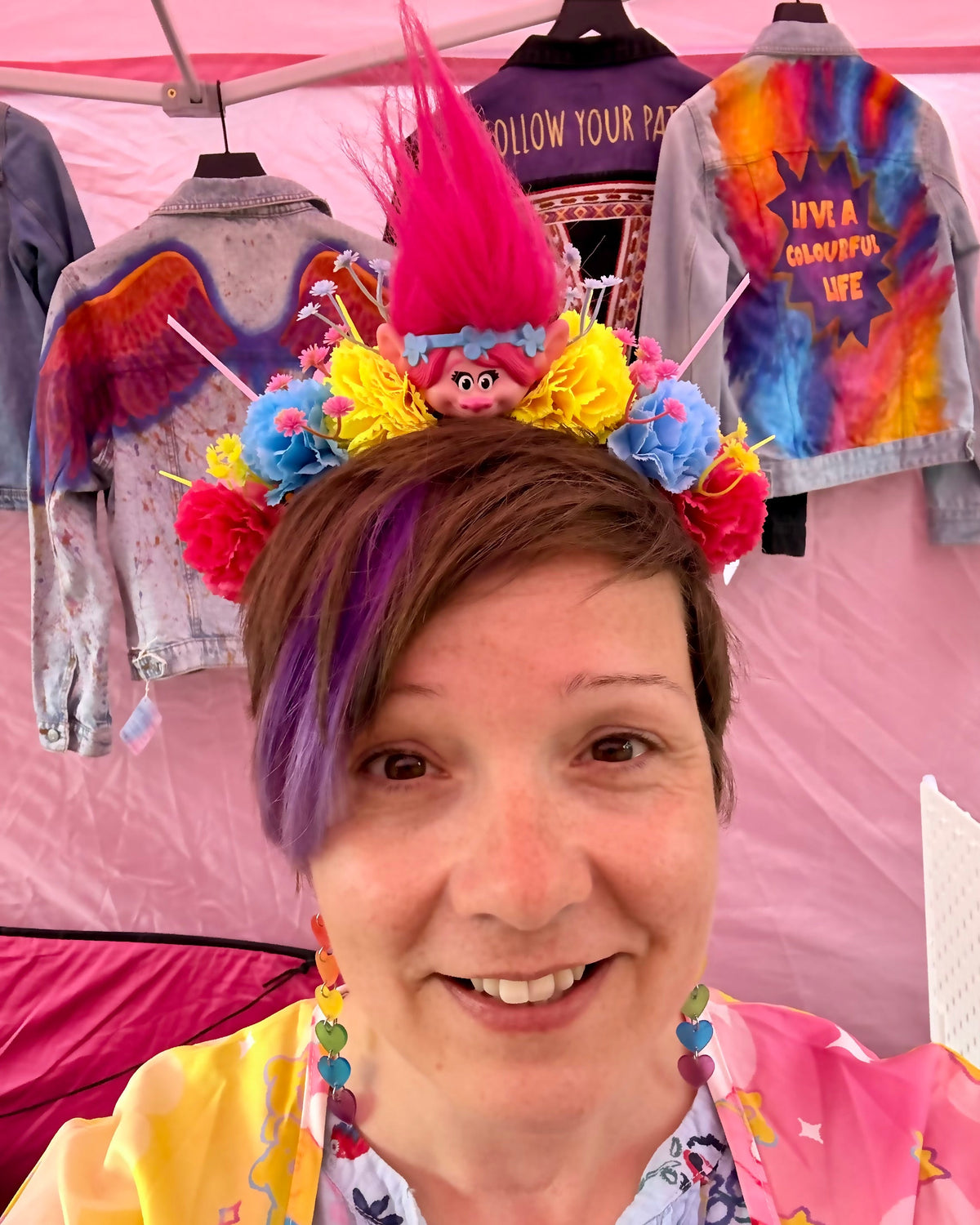 A smiling woman at an outdoor market wearing a colourful Trolls-themed festival crown with a bright pink mohawk, yellow, blue, and pink flowers, and a small character figure. Background shows colourful hand painted denim jackets hanging under a pink canopy.