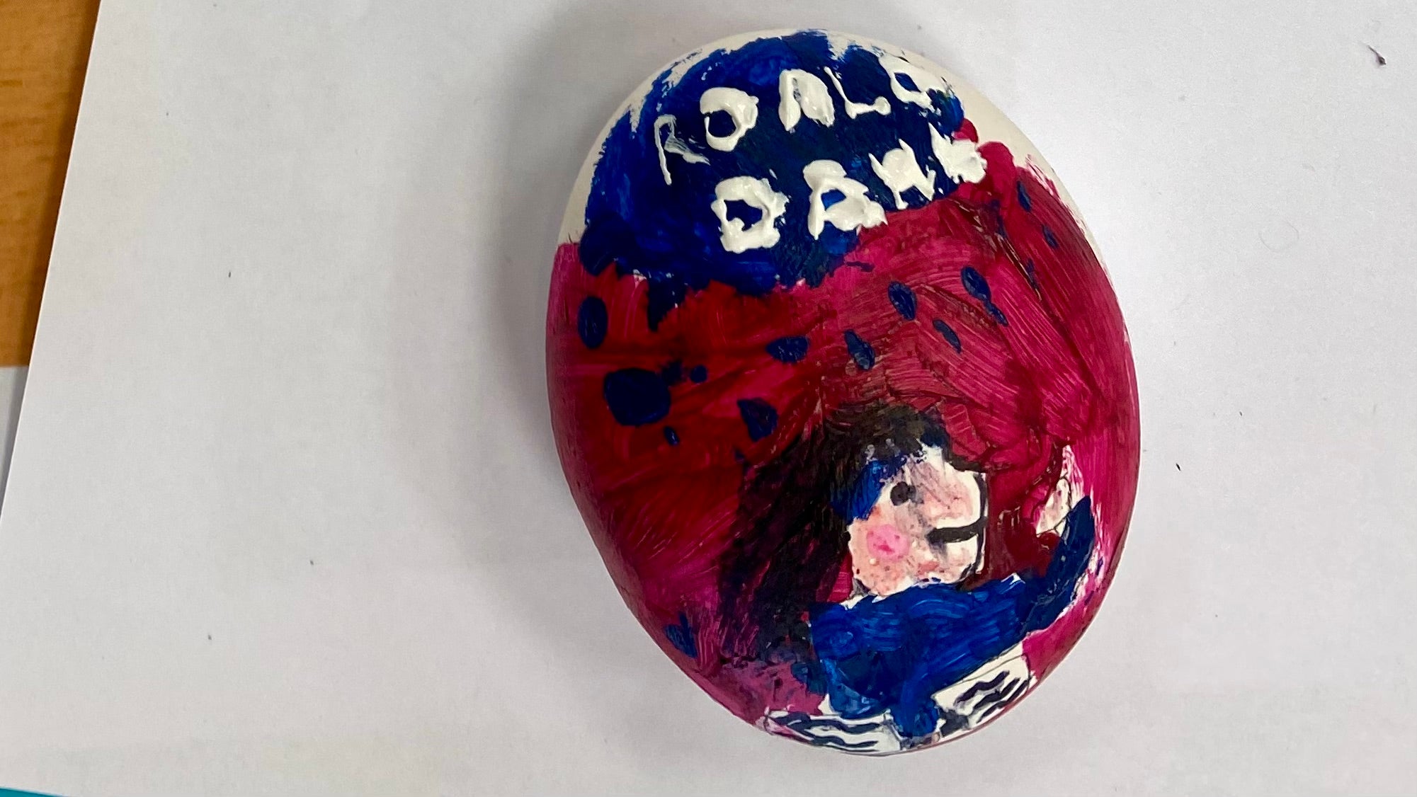 A painted pebble painted to look like the front cover of the book Matilda by Roald Dahl. Reds and blues with a painting of a girl and the words 'Roald Dahl' above.